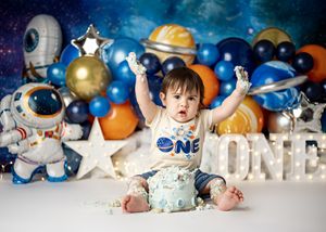 First birthday cake smash photo with space theme decorations including astronauts, stars and blue balloons.