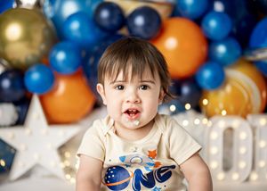 A small child sits in front of a festive backdrop with blue and orange balloons and decorative stars.