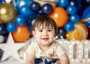 Baby in tan shirt sits against festive backdrop with blue and orange balloons and glowing star decorations.