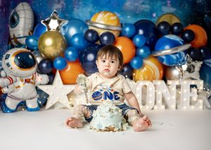 Space themed first birthday cake smash setup with astronaut decorations and blue balloon garland against dark backdrop.