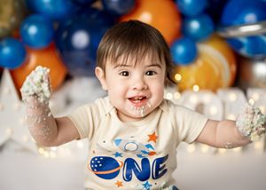 Baby celebrates first birthday with cake smash photo session against blue and orange balloon backdrop.