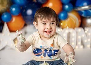 Happy baby wearing a 'ONE' t-shirt celebrates their first birthday with blue and orange balloons in the background.
