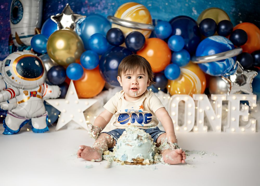 Colorful space themed first birthday cake smash setup with blue and orange balloons, stars, and rockets against blue background.