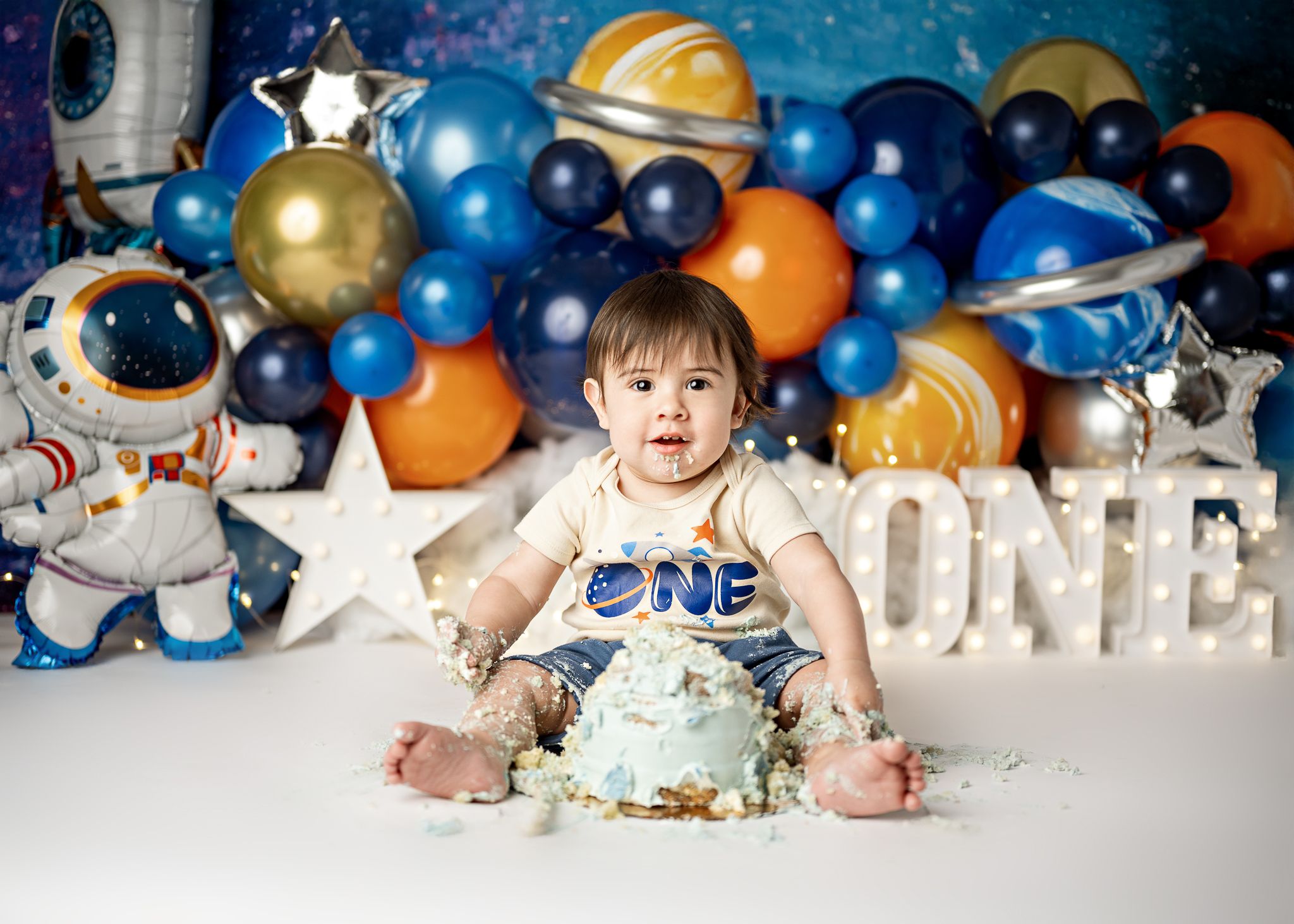 Colorful space themed first birthday cake smash setup with blue and orange balloons, stars, and rockets against blue background.