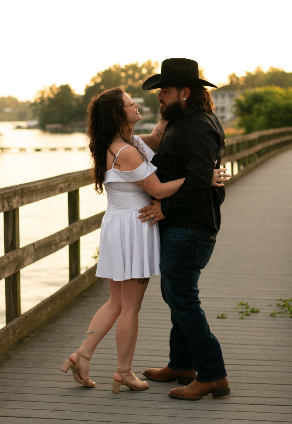 A couple in western attire dances lovingly on a wooden boardwalk at sunset near the water.