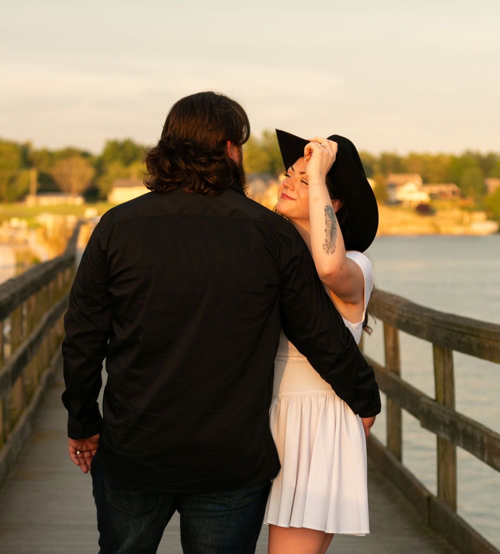 A romantic couple in black and white attire shares an intimate moment on a wooden dock at sunset by the water.