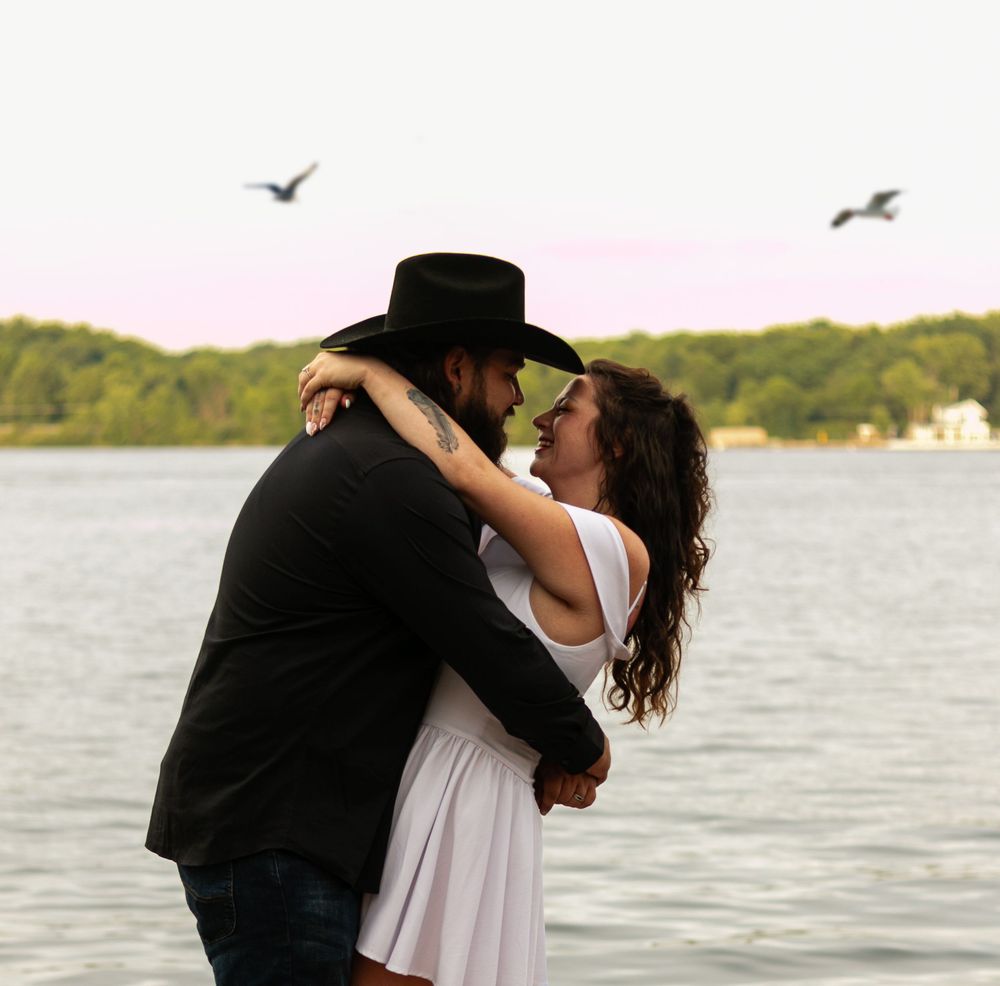 Couple embraced in a romantic dance near a serene lake at sunset with birds flying overhead.