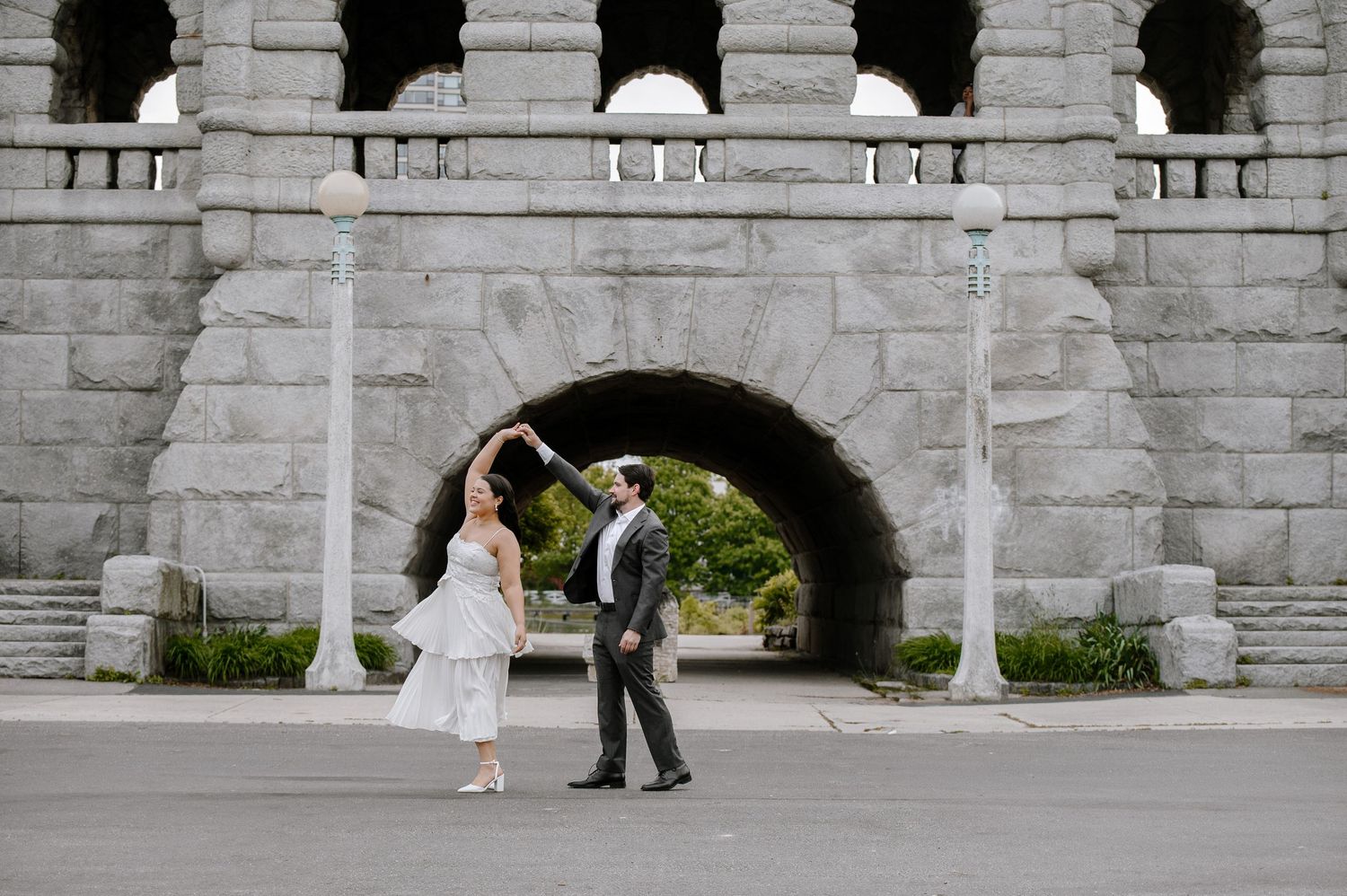 Wedding couple walks through stone archway as photographer captures sequential moments on sunny day.