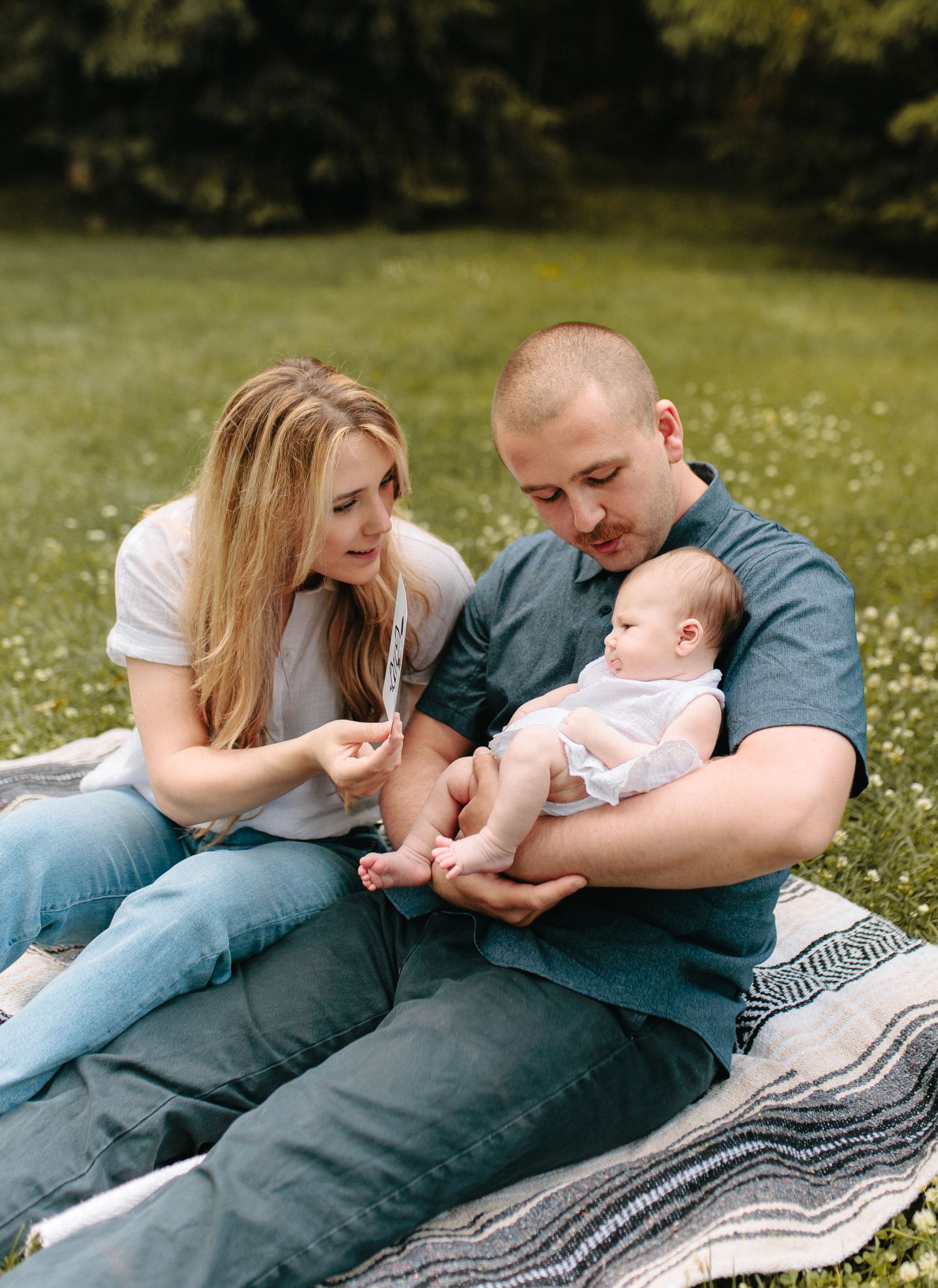 A couple sits on a striped blanket in a grassy field while tenderly caring for their infant on a sunny afternoon.