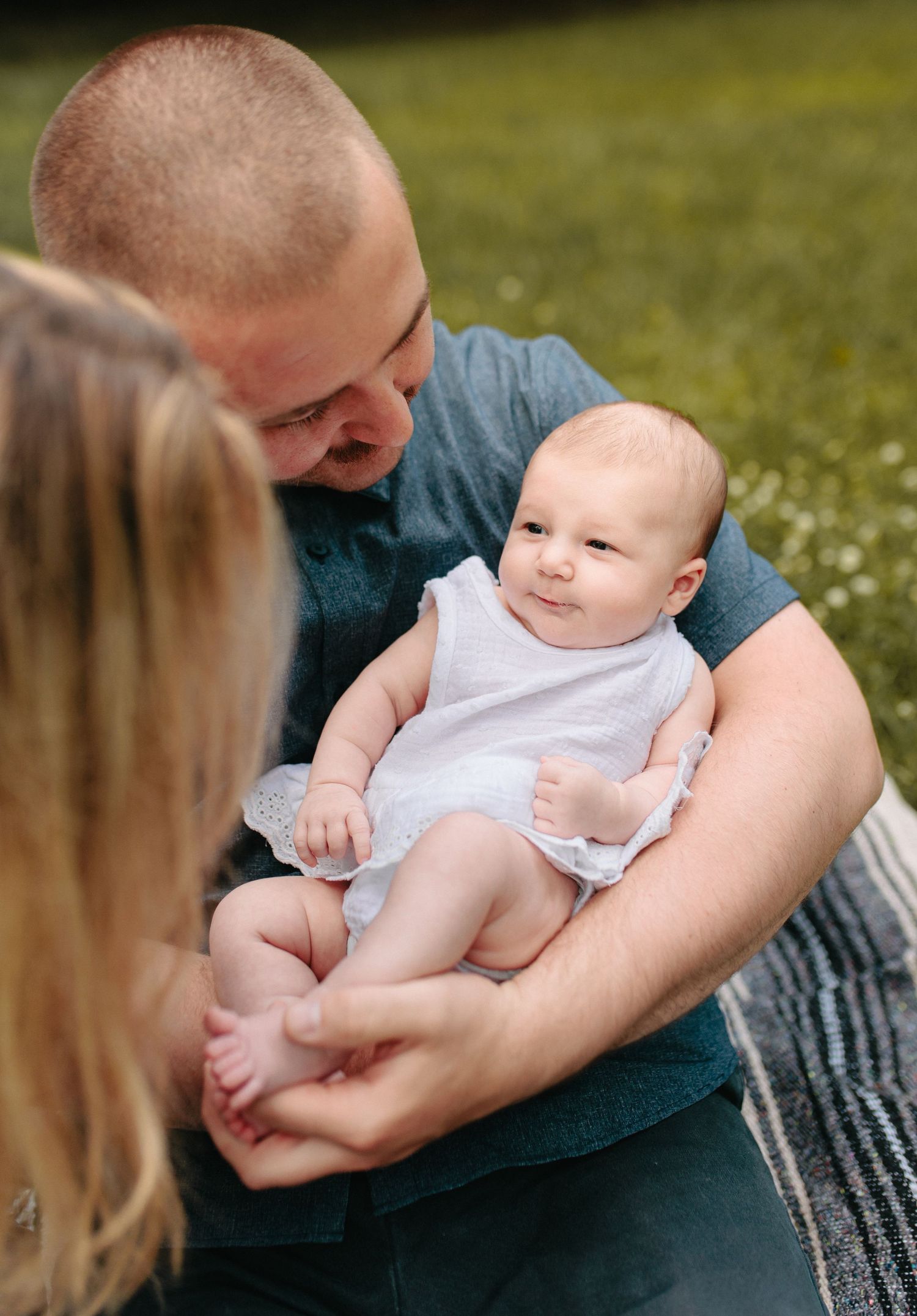A sweet family moment captured outdoors on grass with a smiling baby in a white outfit.