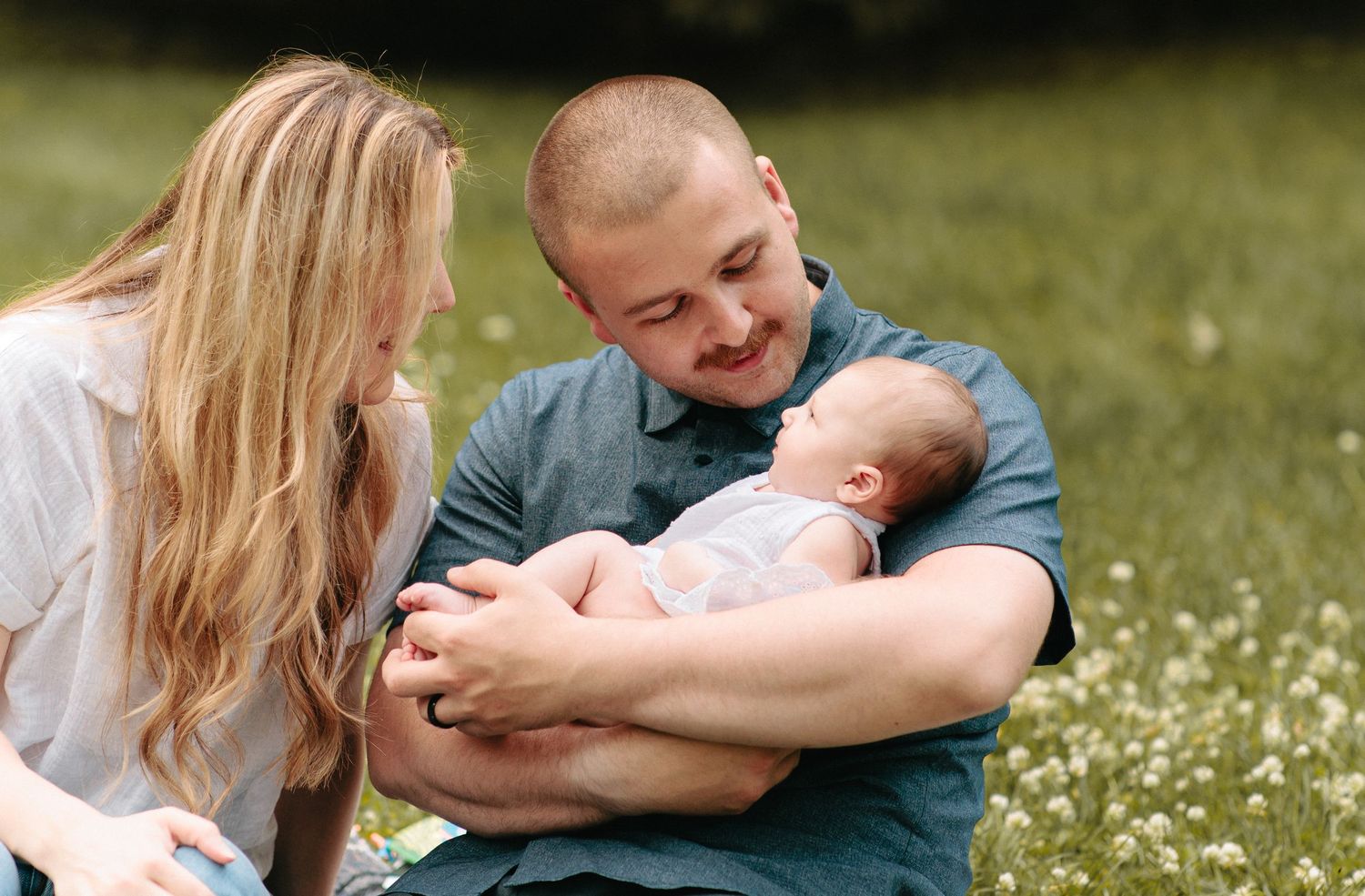 Happy couple sitting in a wildflower meadow holding their sleeping newborn baby on a sunny day.