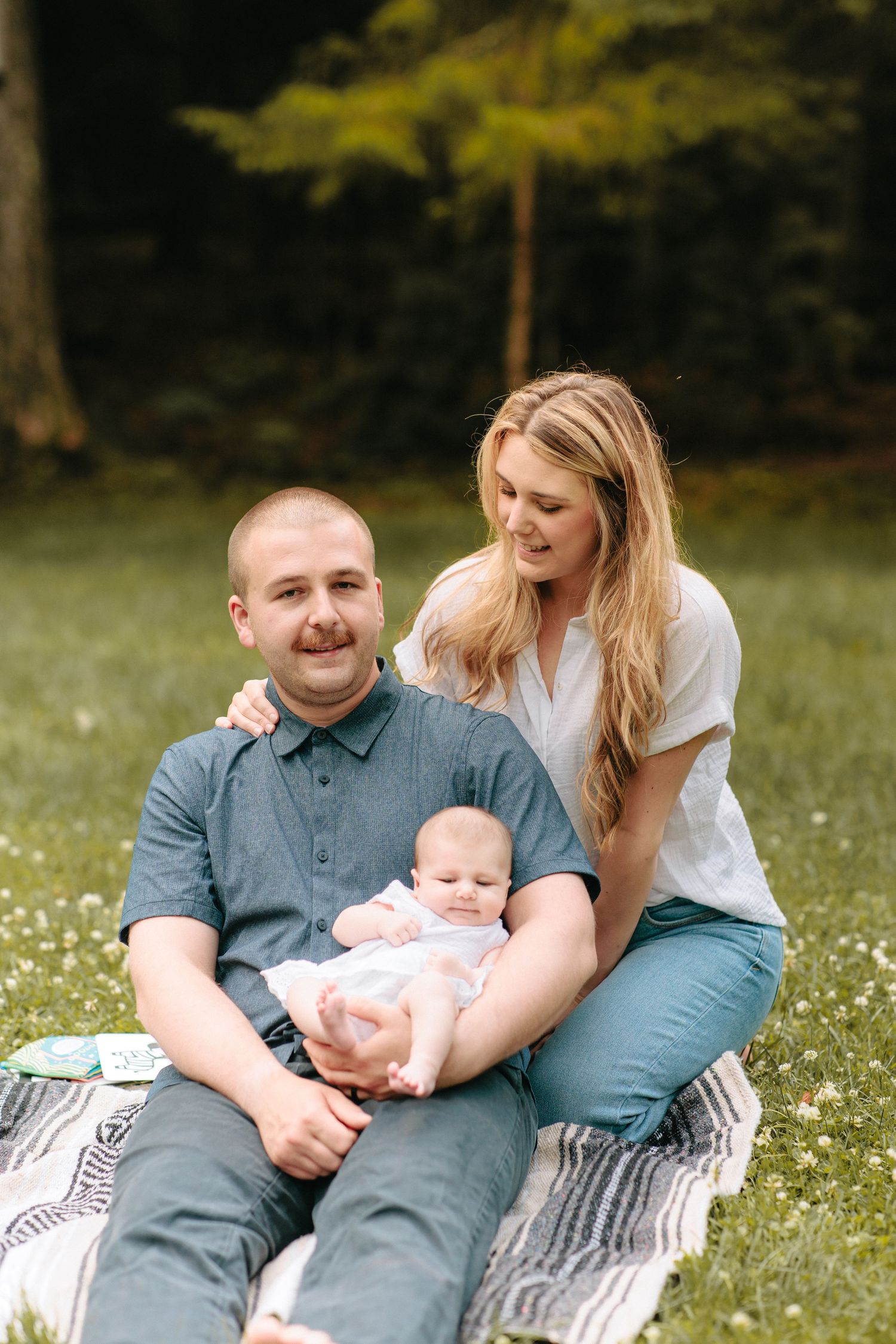 A family sits together on a striped blanket in a grassy field during a sunny outdoor photo session.