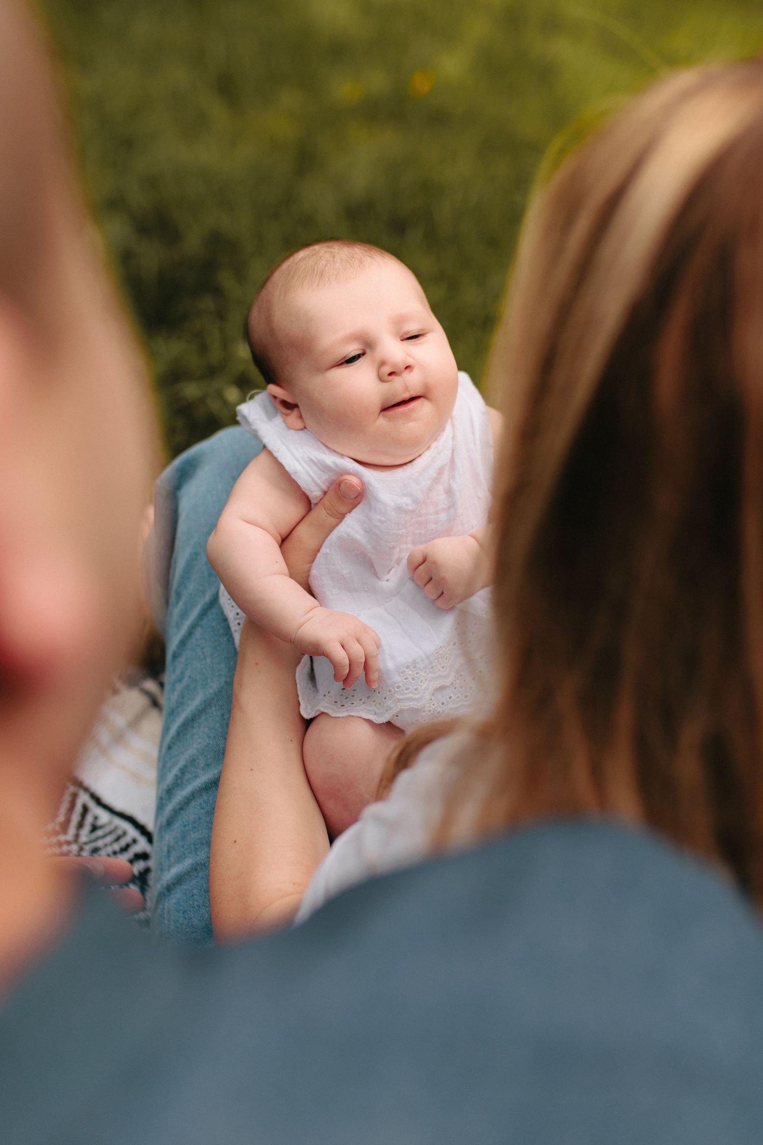 A small baby in a white romper smiles while being held outdoors on a sunny day.