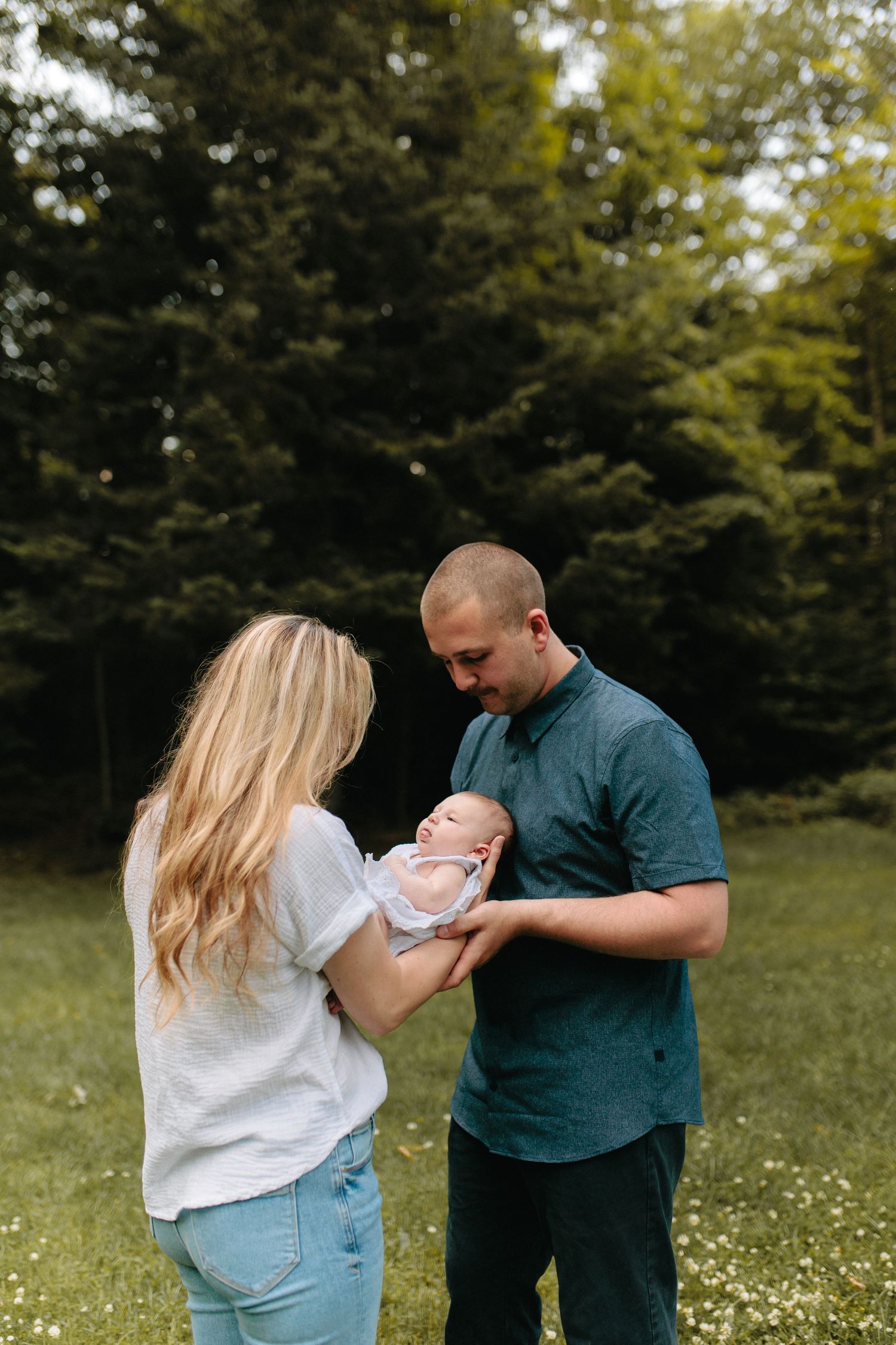 A couple shares a tender moment with their baby while standing in a wooded area with wildflowers.