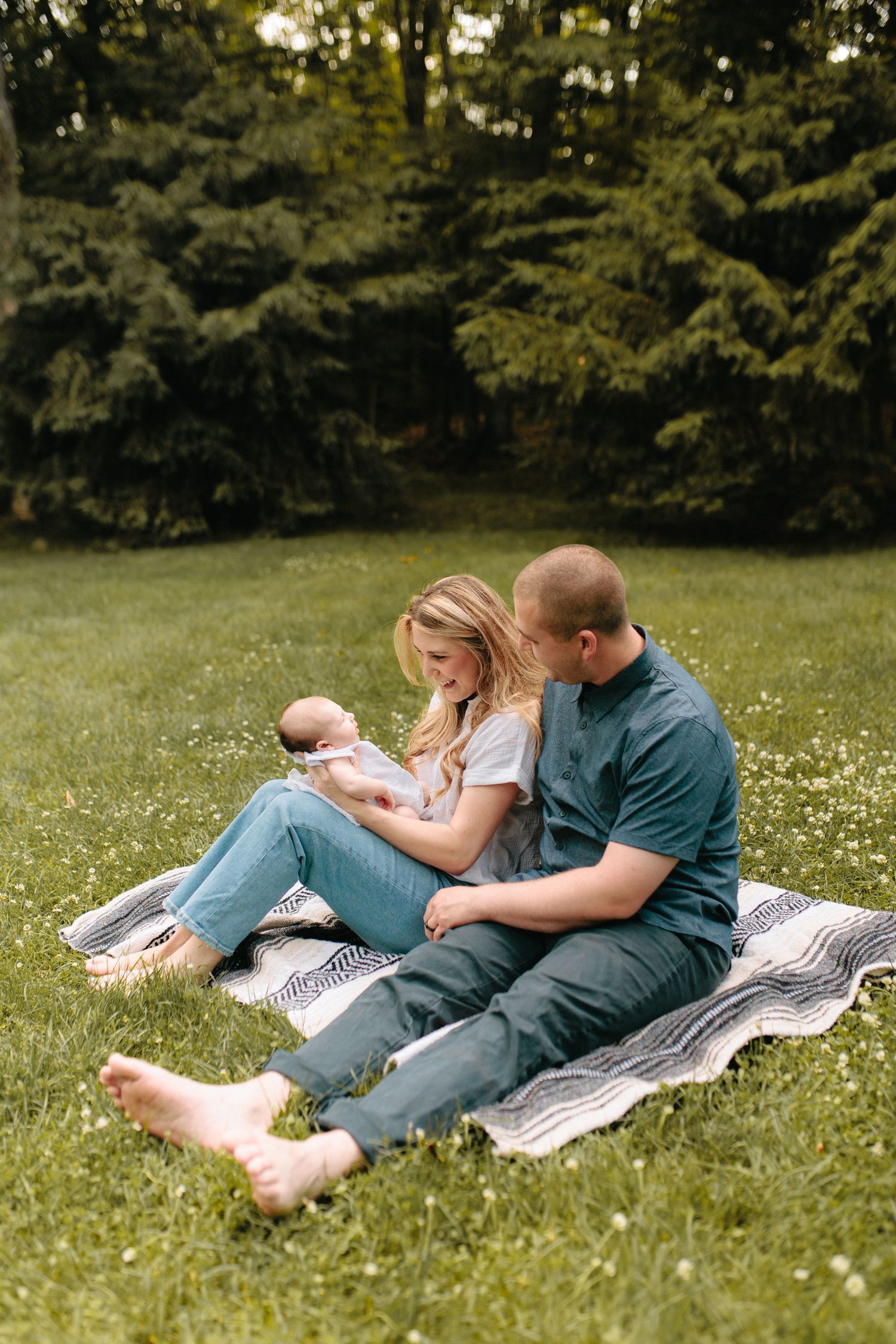 Family relaxes on picnic blanket with newborn baby in grassy backyard with evergreen trees in background.
