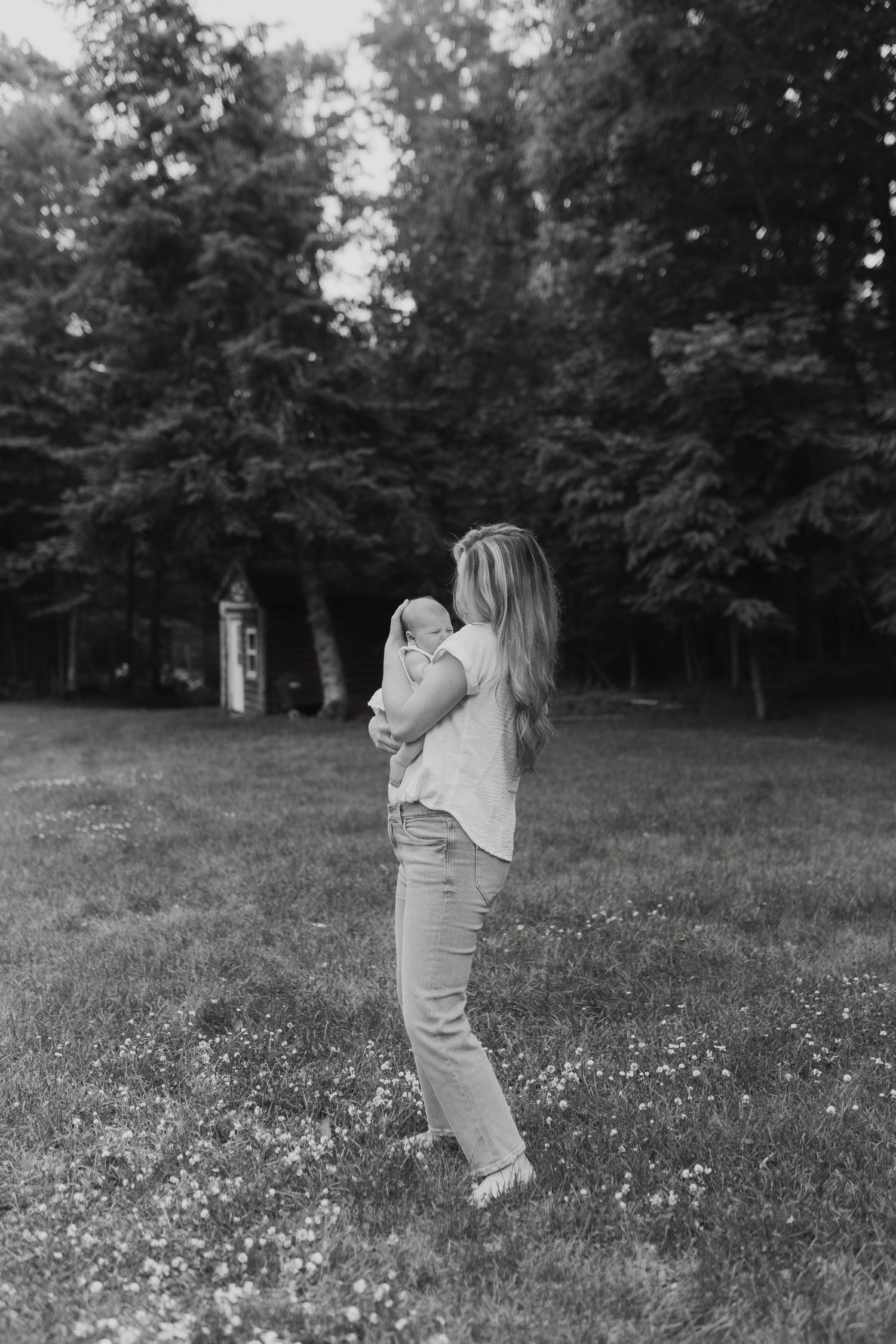 A tender moment captured in black and white as a caregiver holds a baby while standing in a meadow with trees in background.