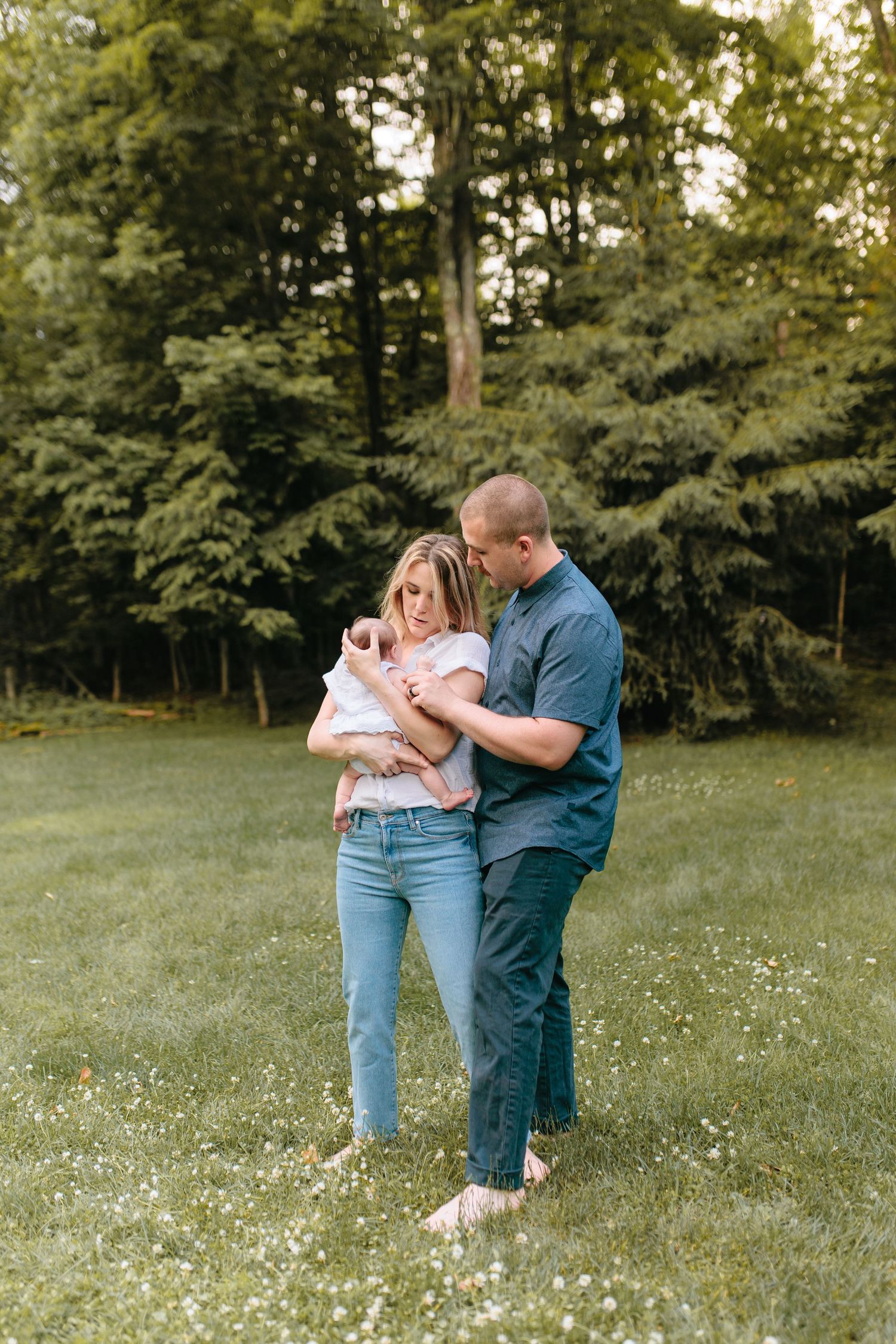 A couple stands together holding a baby in a grassy field surrounded by lush green trees on a sunny day.