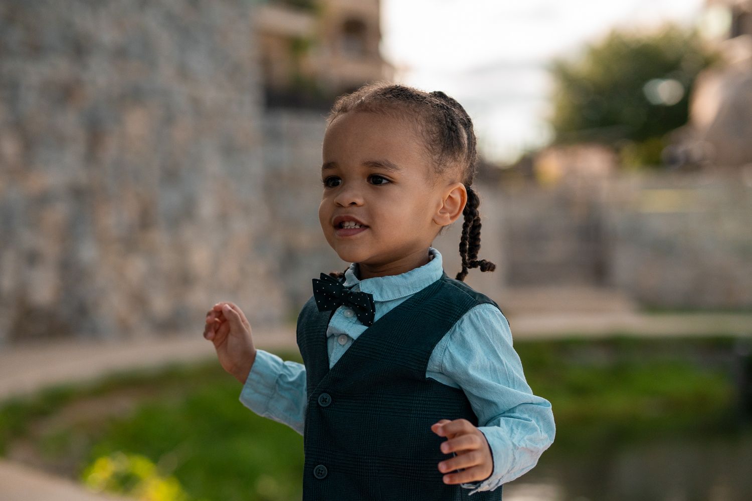Toddler in a formal vest and bow tie smiles while standing outdoors in late afternoon sunlight.