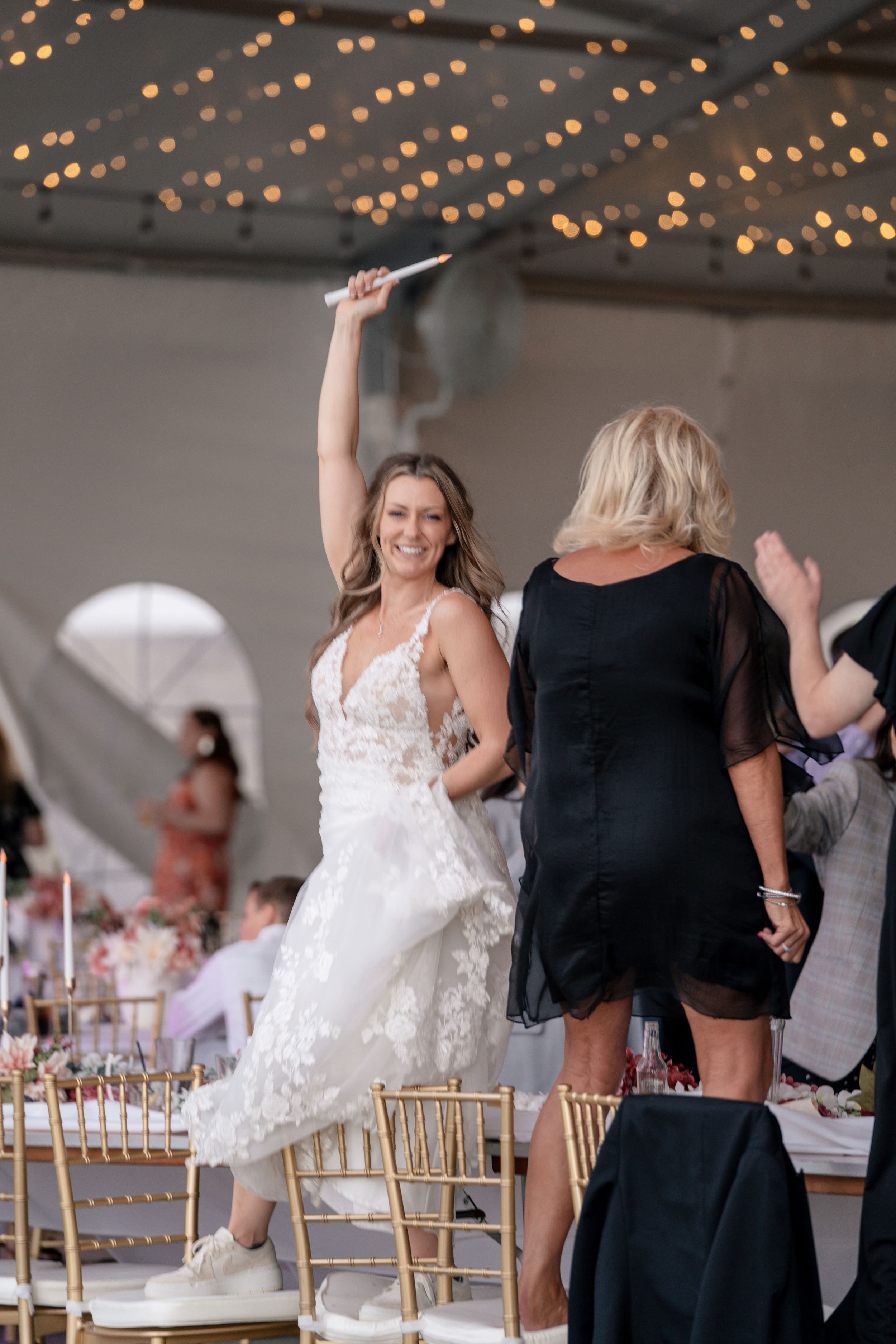 Bride in white lace wedding dress and sneakers dances joyfully at reception under string lights with gold chairs visible.