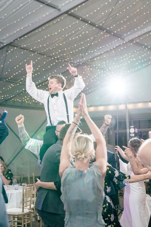Wedding guests dance and celebrate with raised arms under a string-lit tent ceiling during a reception.