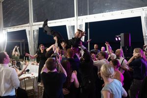 A lively crowd dances and celebrates during an evening reception with purple lighting in a tented venue.