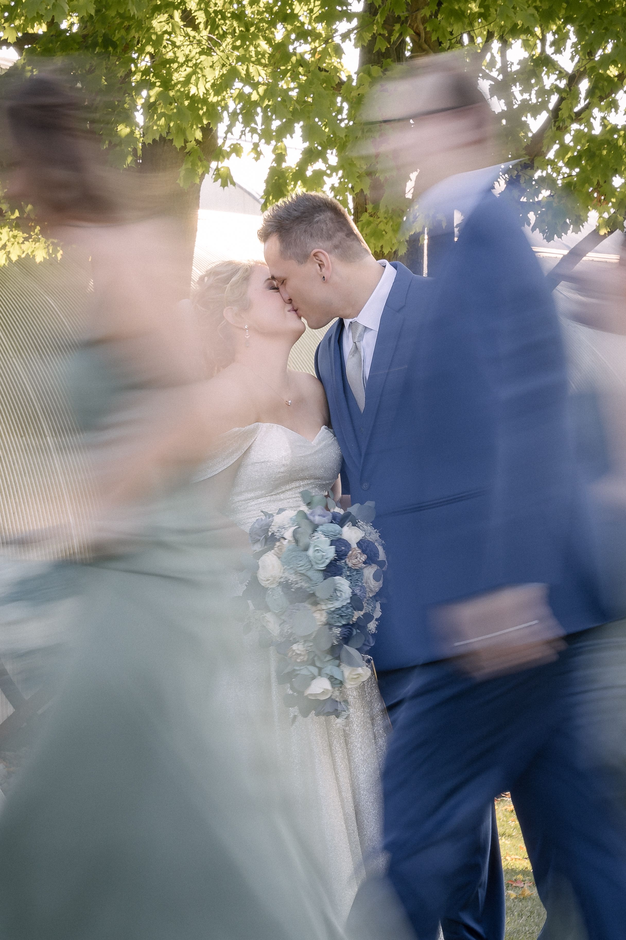 Wedding couple shares a sweet romantic moment under sunlit trees with a blue and white floral bouquet.