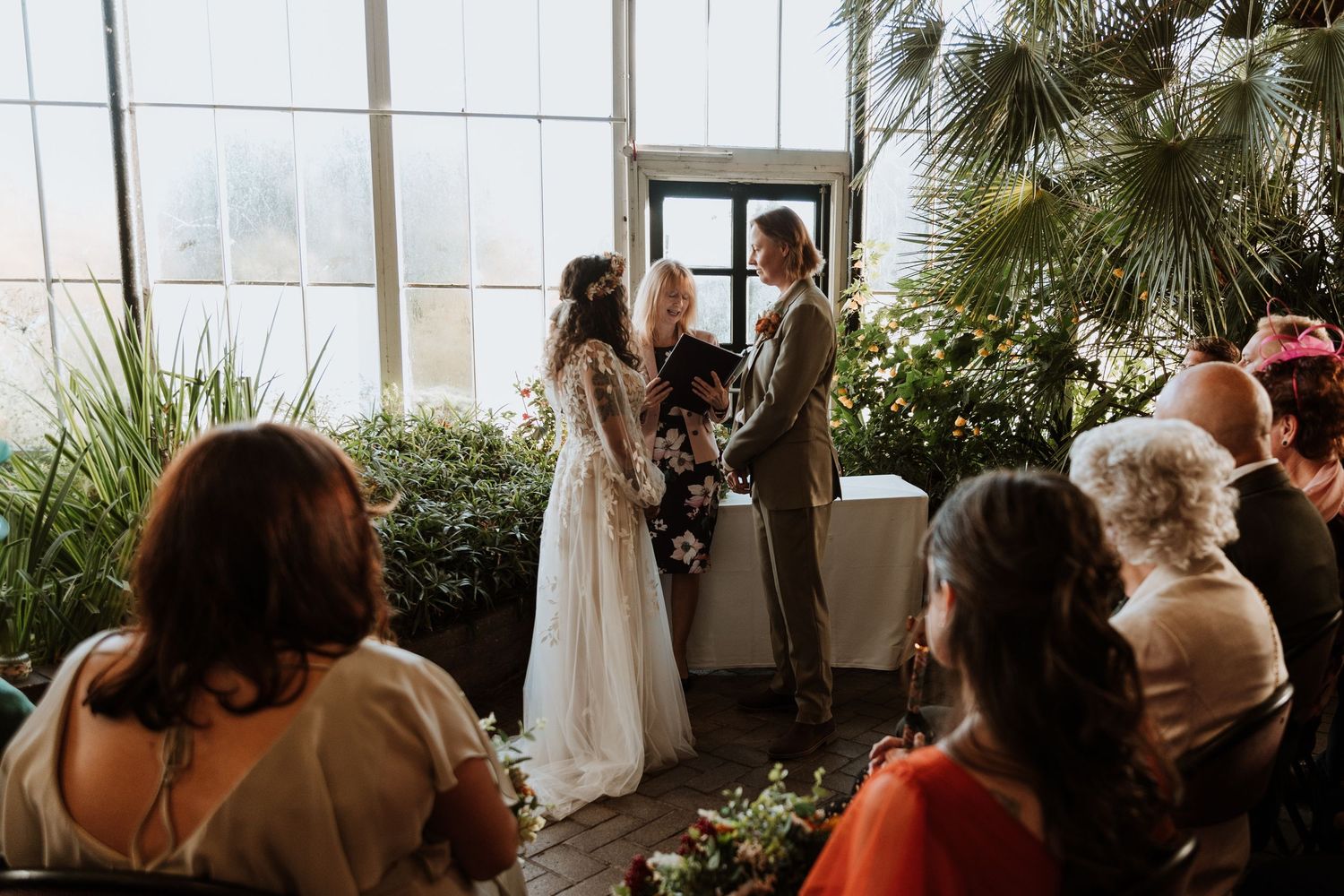 A couple exchanges vows during an indoor greenhouse wedding ceremony surrounded by lush plants and seated guests.