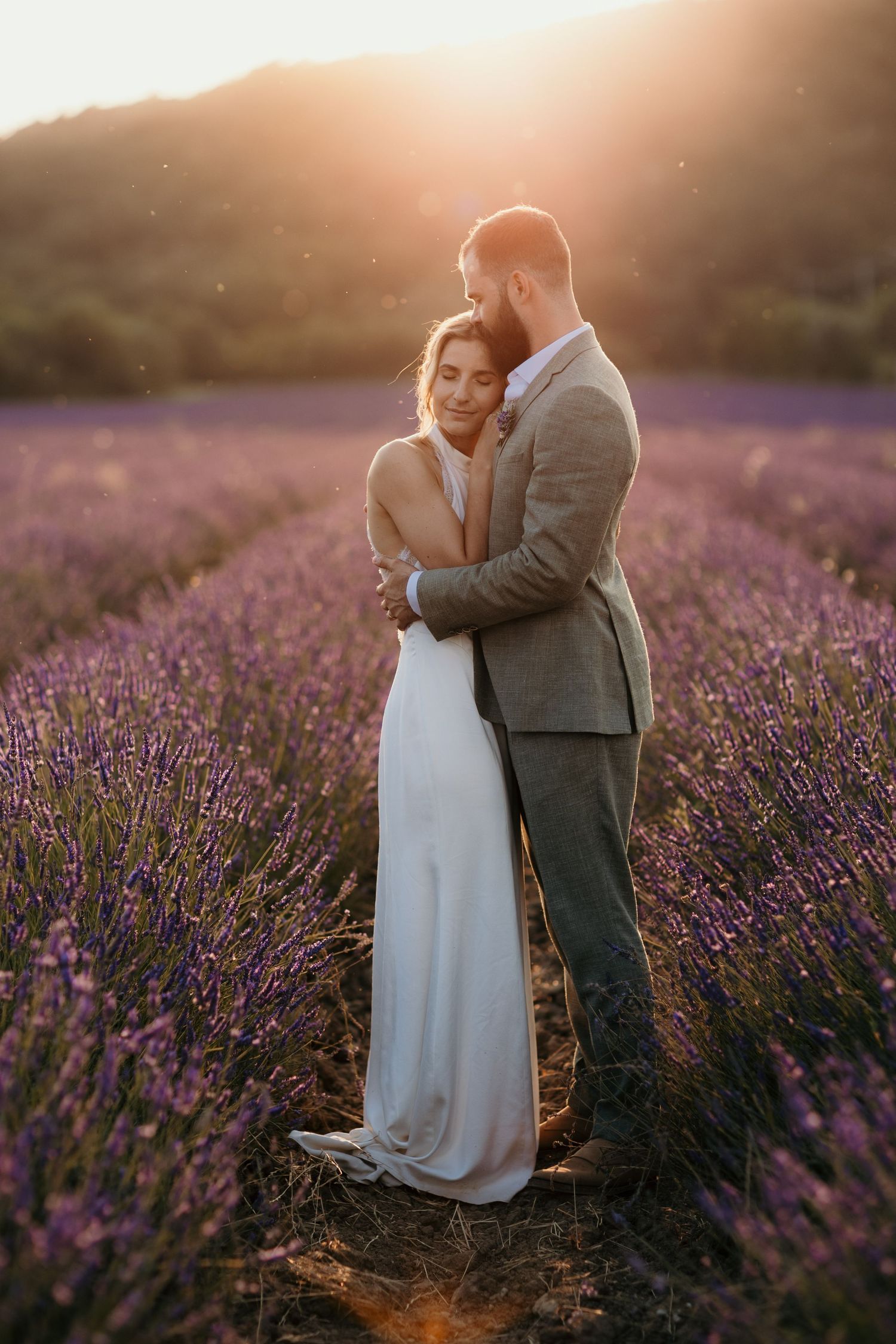 A romantic couple embraces in a sunlit lavender field at sunset wearing wedding attire.