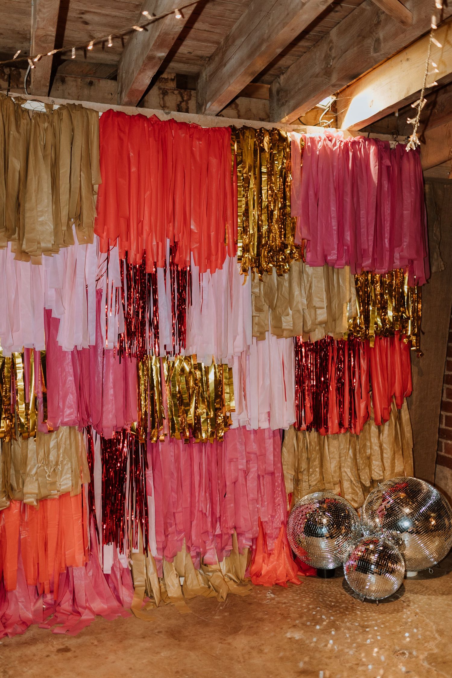 Cascading streamers in pink, coral, gold and red create a festive backdrop wall with disco balls on the floor.