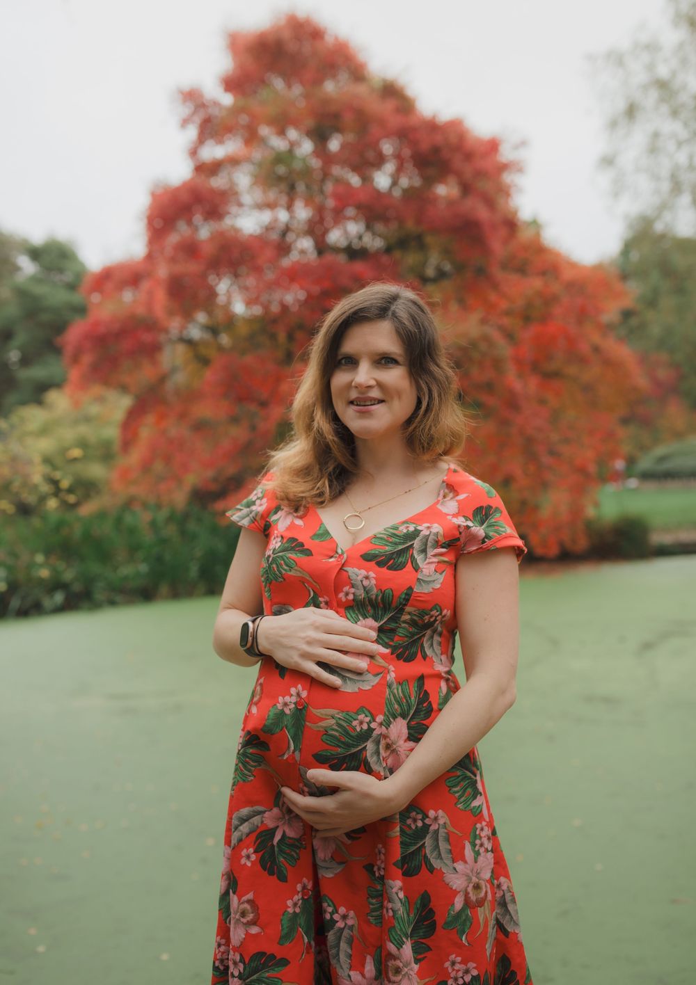 Person in floral red dress stands against beautiful fall maple trees in outdoor setting.