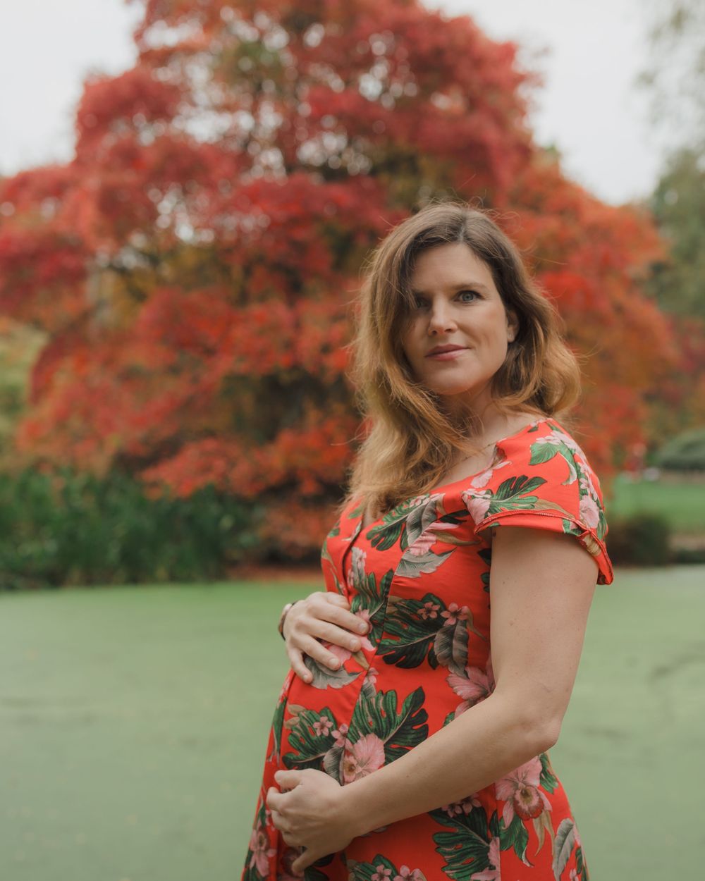 Woman in a red floral dress poses with autumn foliage and colorful trees in the background.