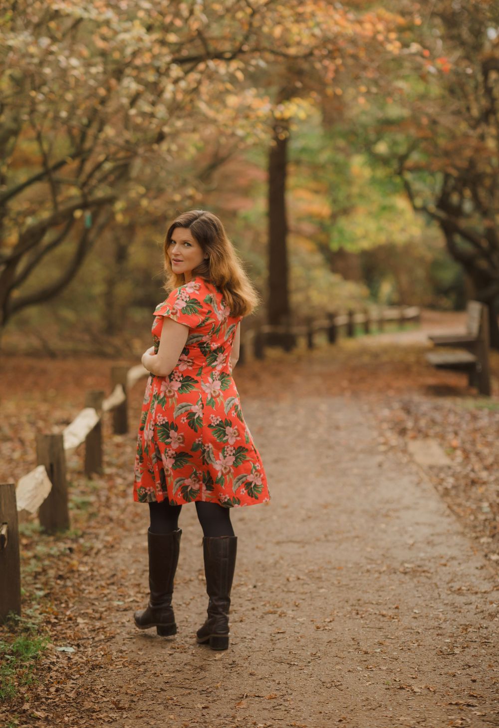 A person in a red floral top and boots stands on a leaf-covered autumn path in a park.