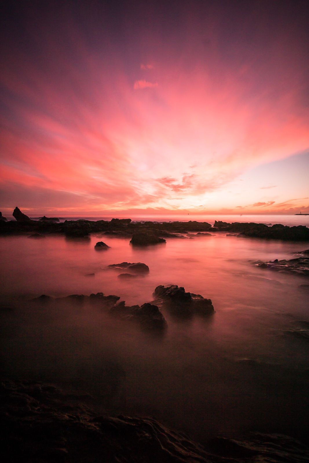 Dramatic pink sunset illuminates rocky coastline with silhouetted rocks emerging from misty ocean waters under vibrant sky.