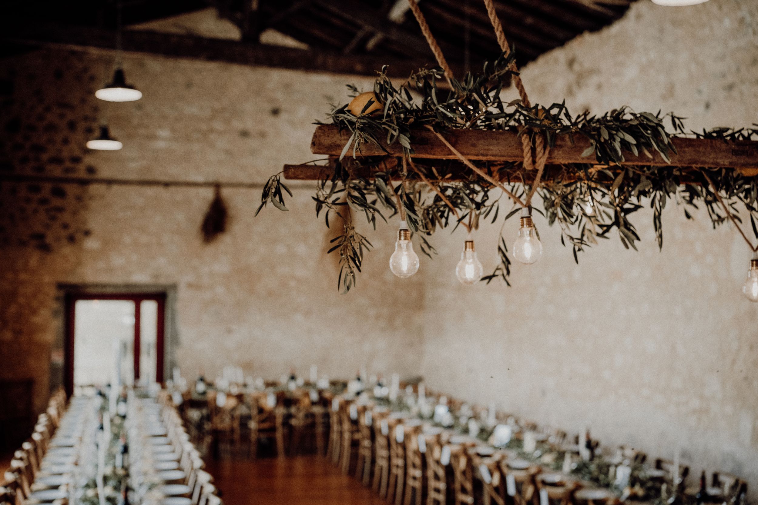 Wedding venue interior with long dining tables and hanging greenery decor from rustic wooden beams.