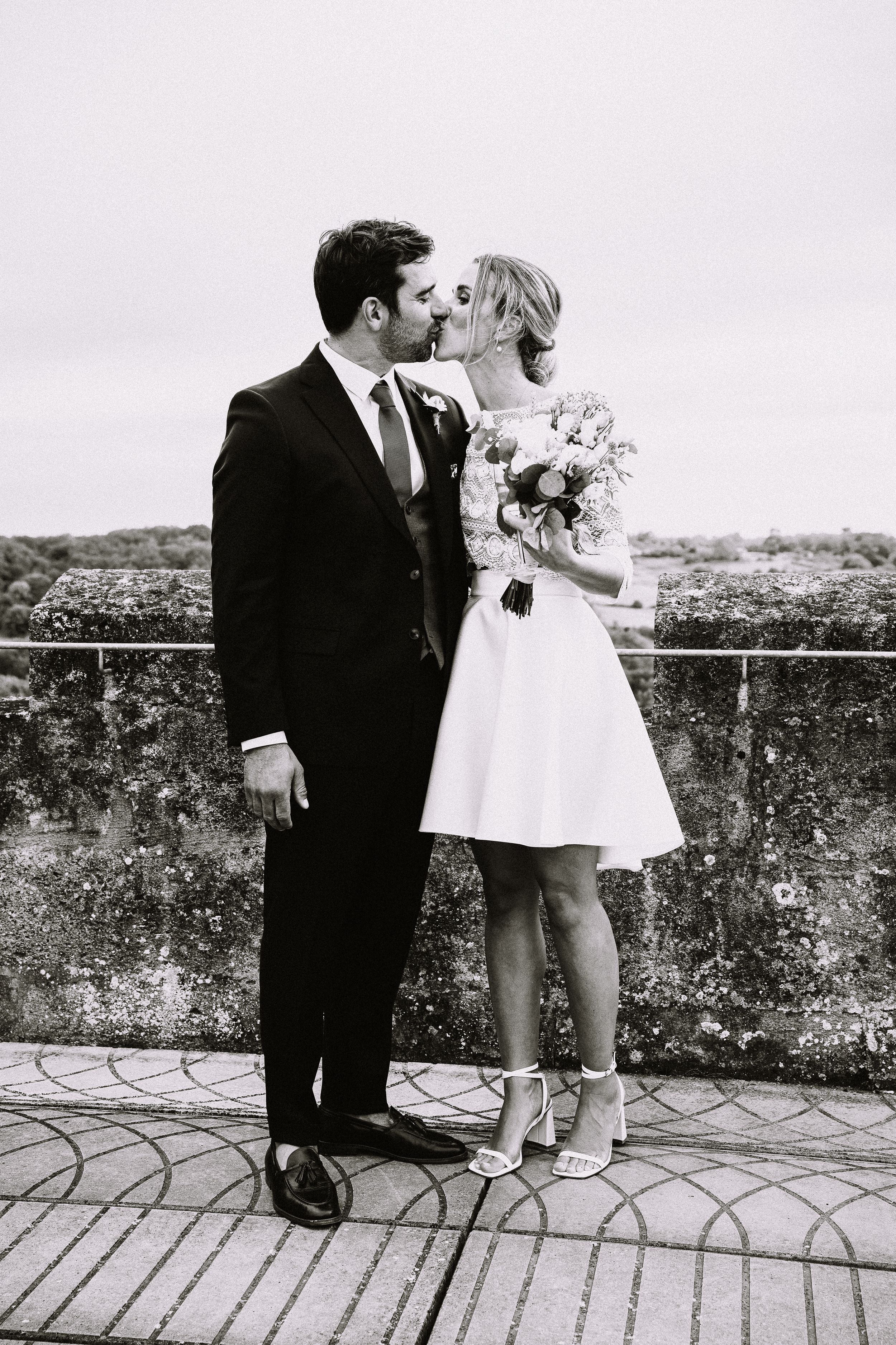 Black and white wedding photo of newlyweds sharing an intimate moment on a stone balcony with bouquet.