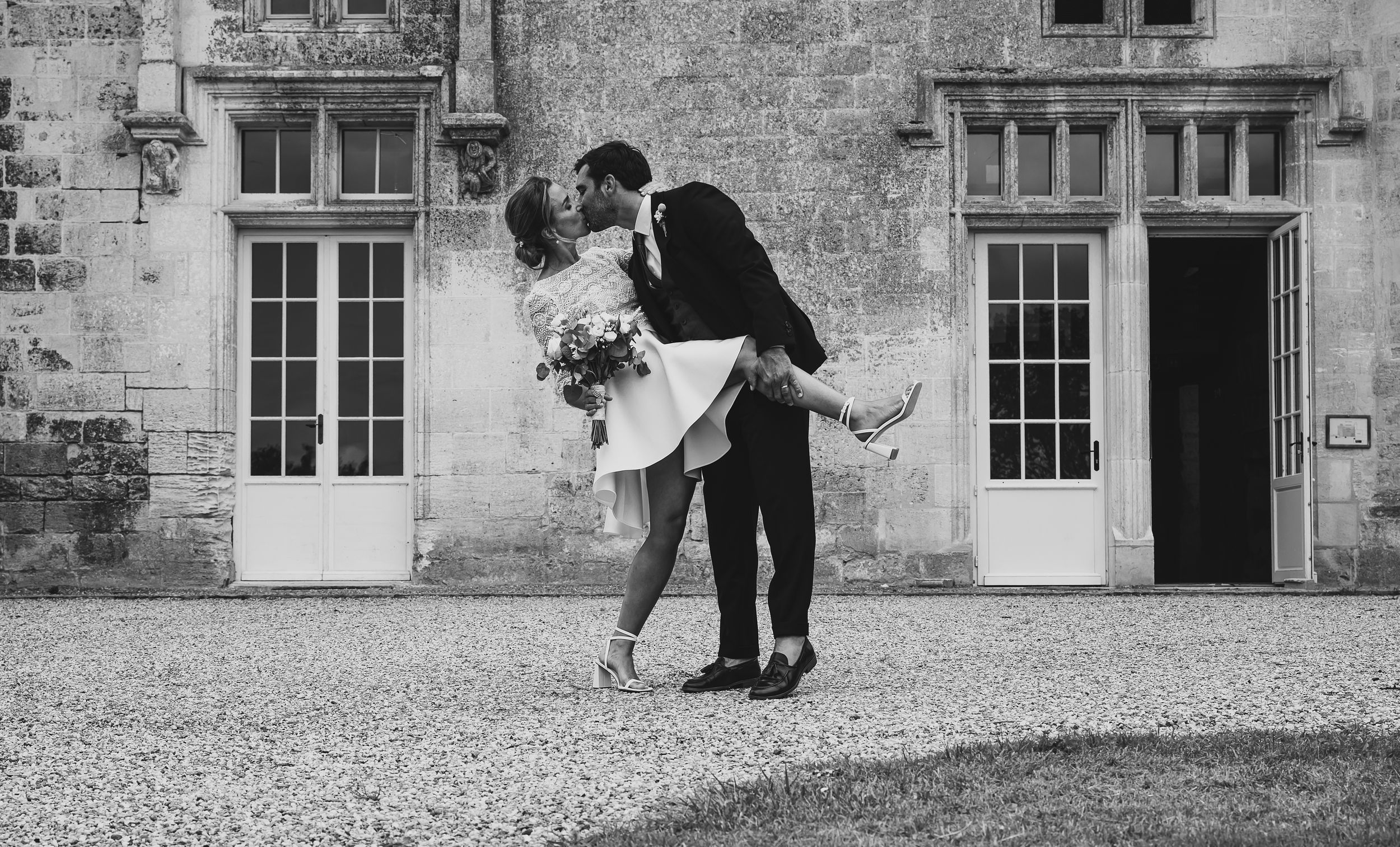 A couple dips and embraces in formal attire on a vintage stone patio with French windows in black and white.