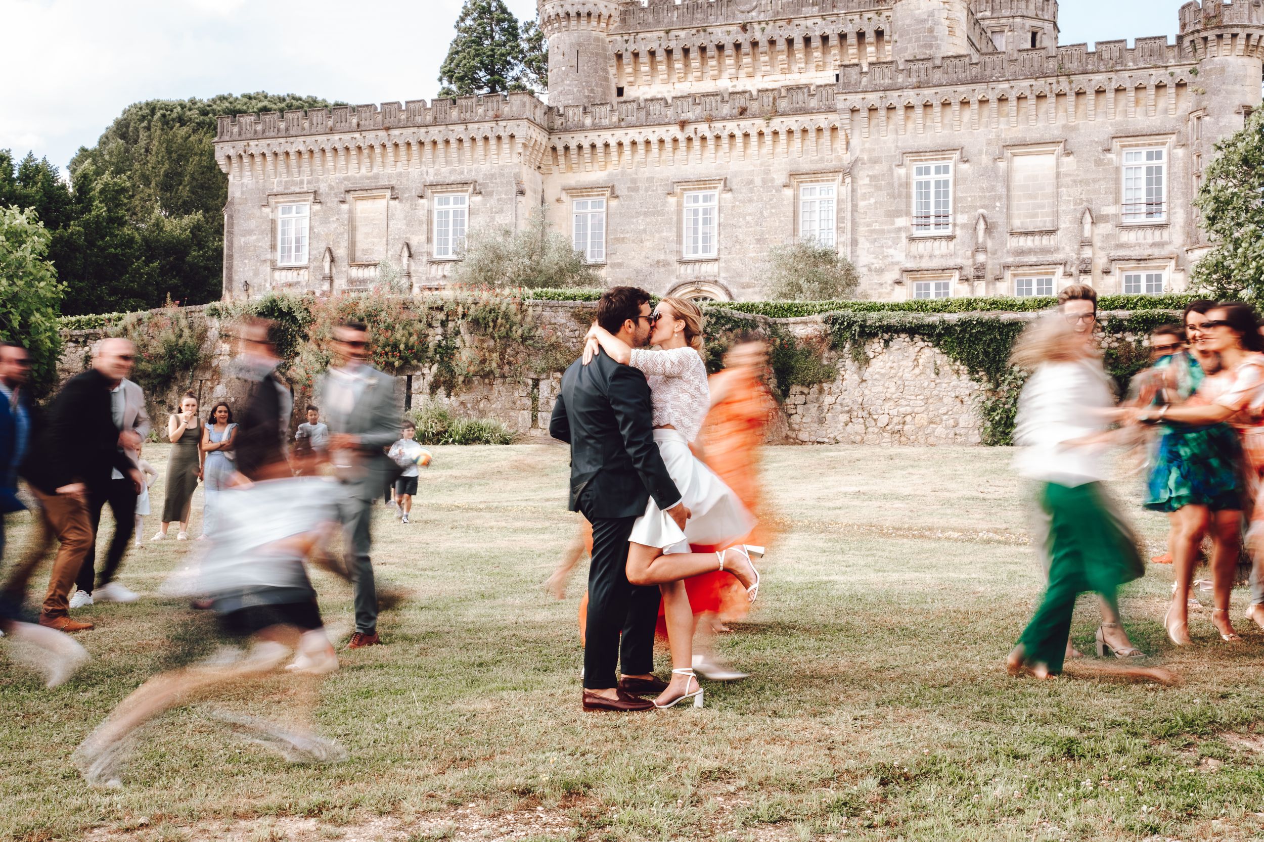 People playing and running on grass outside a historic stone castle with blurred motion capturing festive movement.