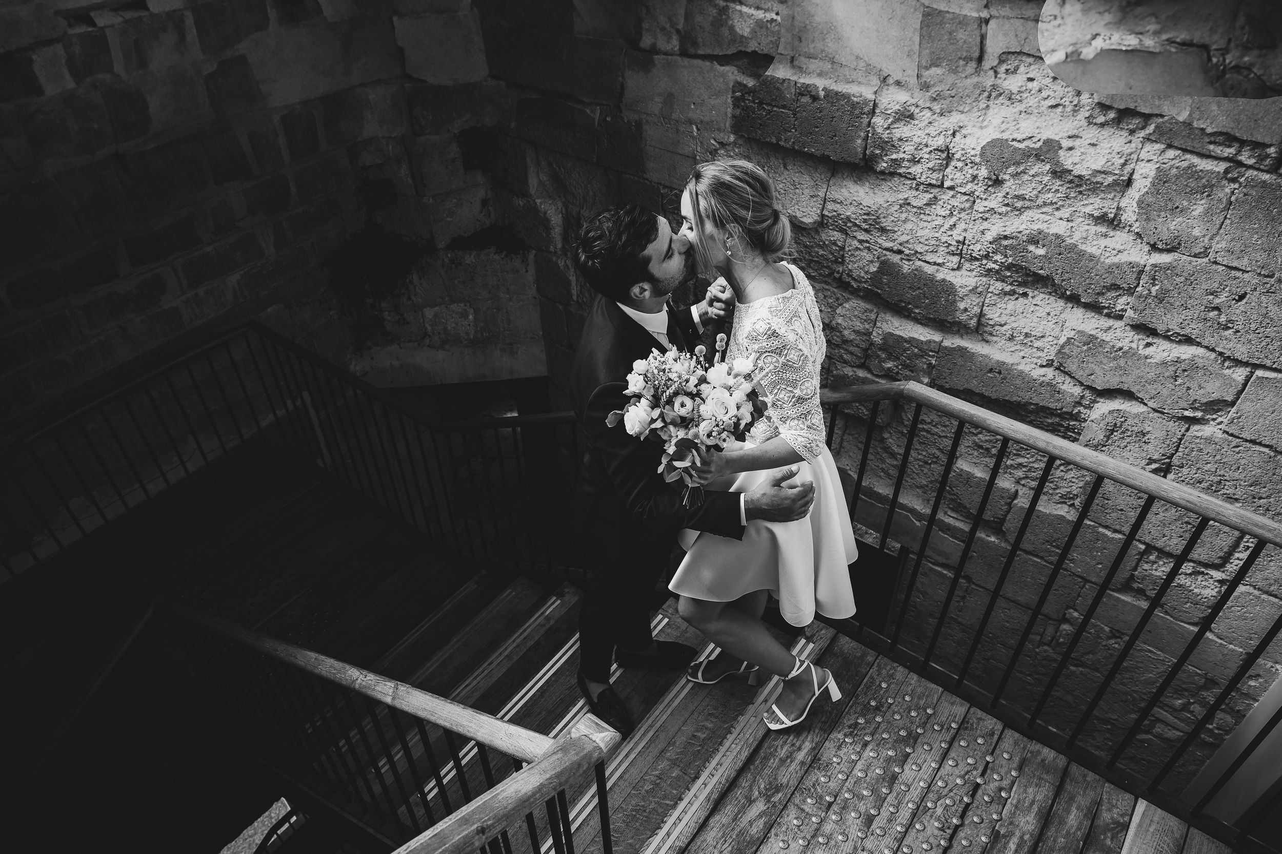 Black and white photo of a couple in winter coats embracing in a rustic stairwell with brick walls.