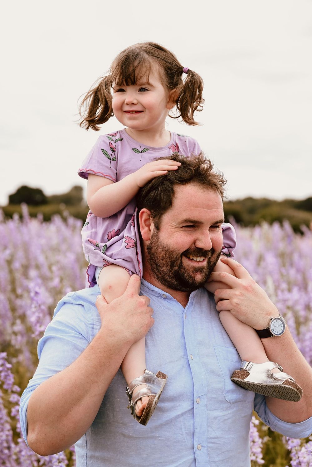 A heartwarming moment captured in a blooming purple wildflower field on a sunny day with a daughter on her dad's shoulders