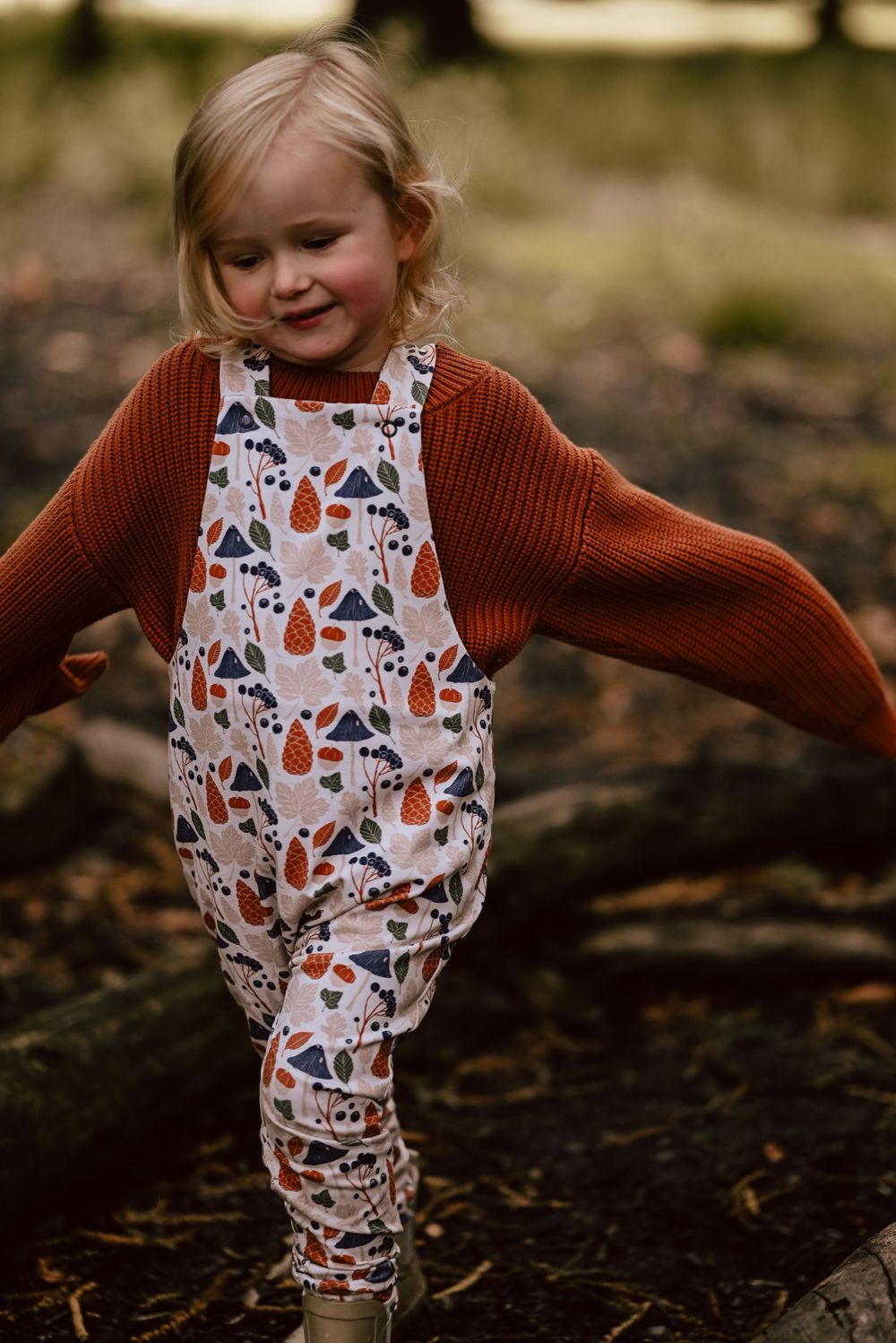 A child wearing a patterned fall-themed overalls and rust-colored knit cardigan plays outdoors on dark soil.