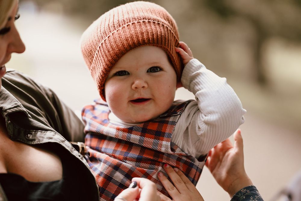Baby wearing a peach knit beanie smiles while being held against blurred outdoor background.