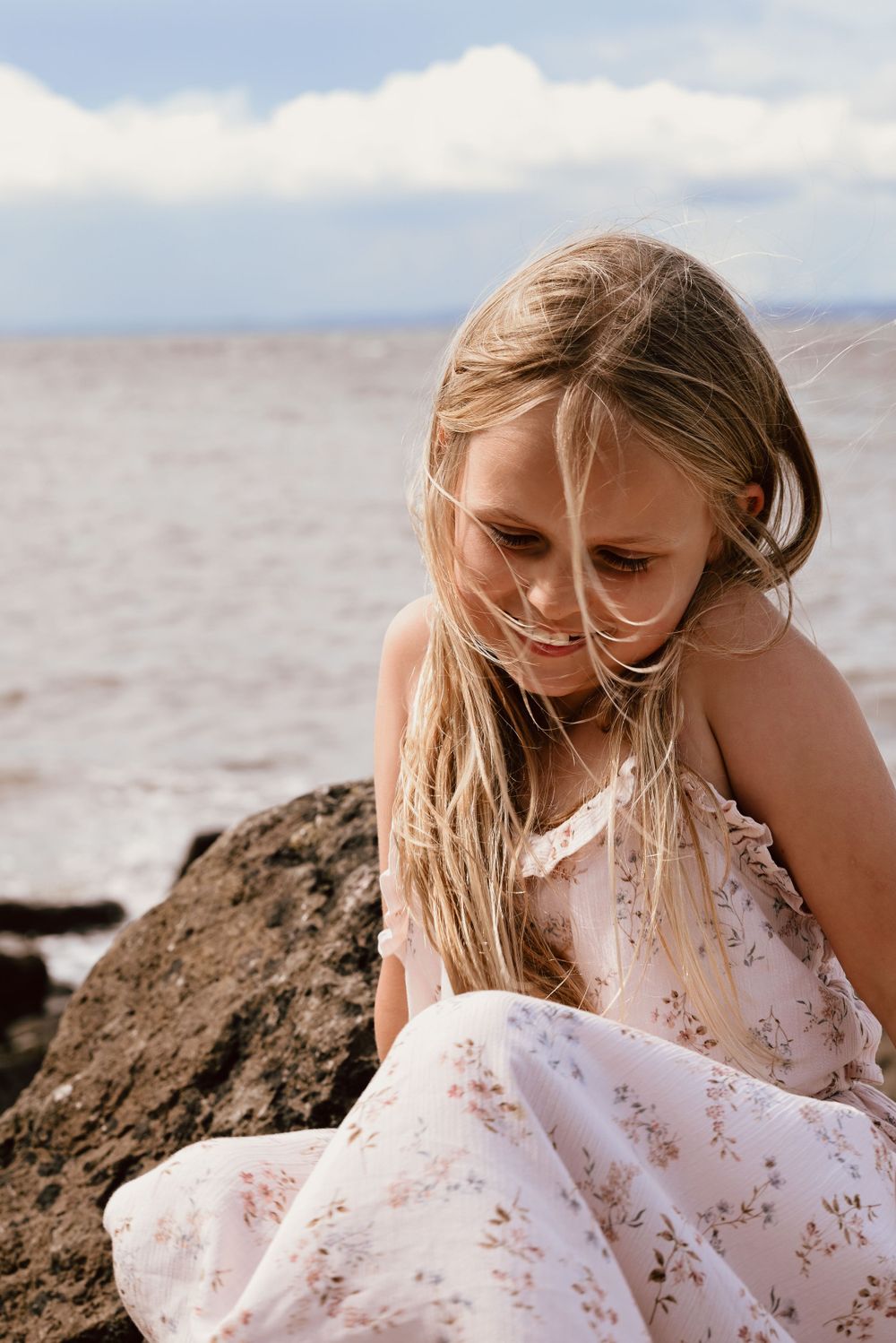 Young blonde girl in floral sundress sits thoughtfully on rocky beach terrain against cloudy sky in Somerset.