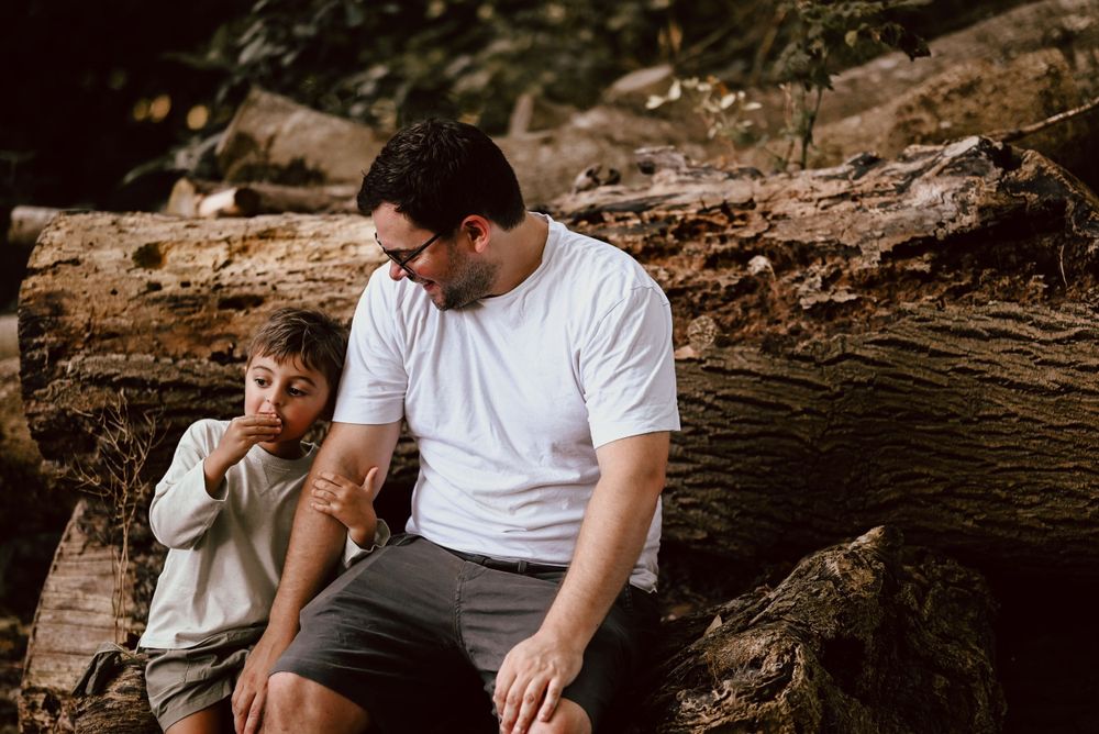 Two people share a tender moment while sitting on a fallen tree trunk in a woodsy outdoor setting.
