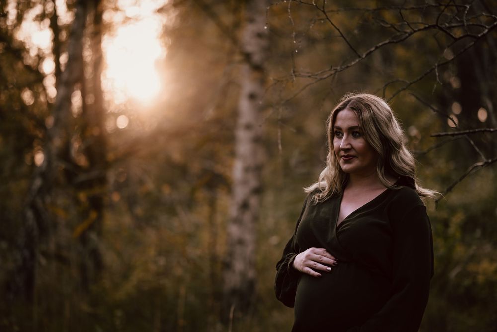 A contemplative pregnant woman stands in a forest at sunset with golden light filtering through birch trees during maternity photoshoot