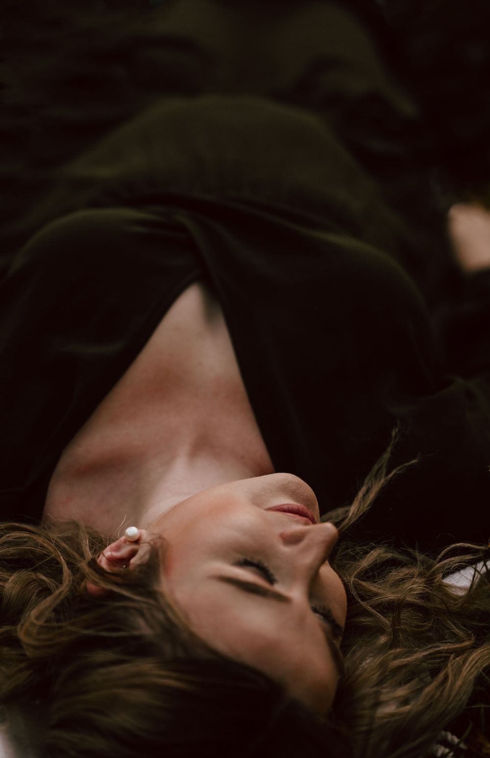 Artistic portrait shows person lying back against dark fabric with soft lighting and scattered hair.
