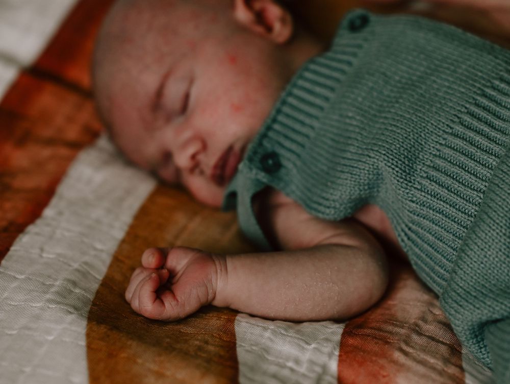 Sleeping infant wearing a green knitted romper lays peacefully on white fabric.
