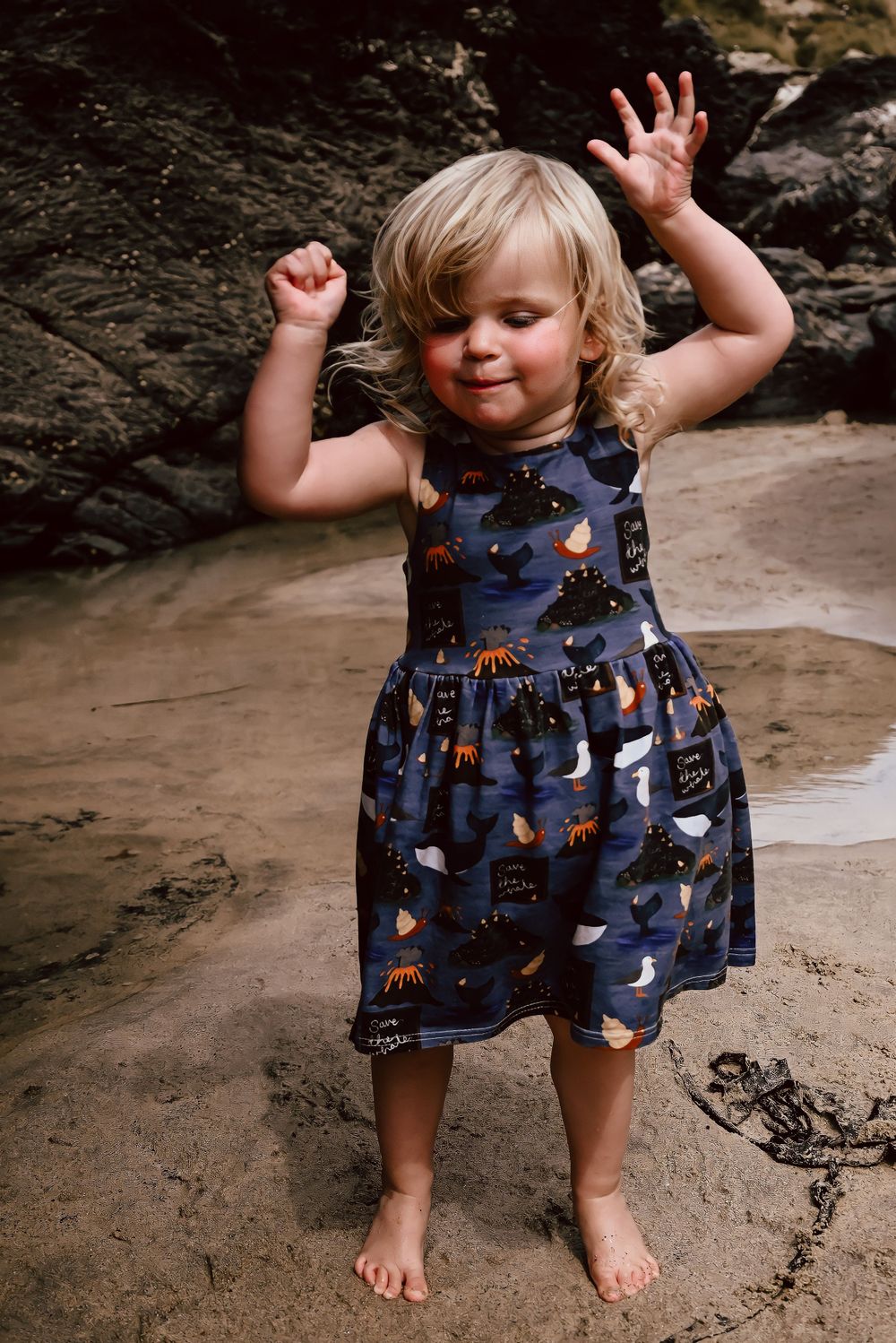 A barefoot child wearing a charming  dress with seaside-themed prints dances joyfully on sandy ground near rocks at Godrevy in Cornwall