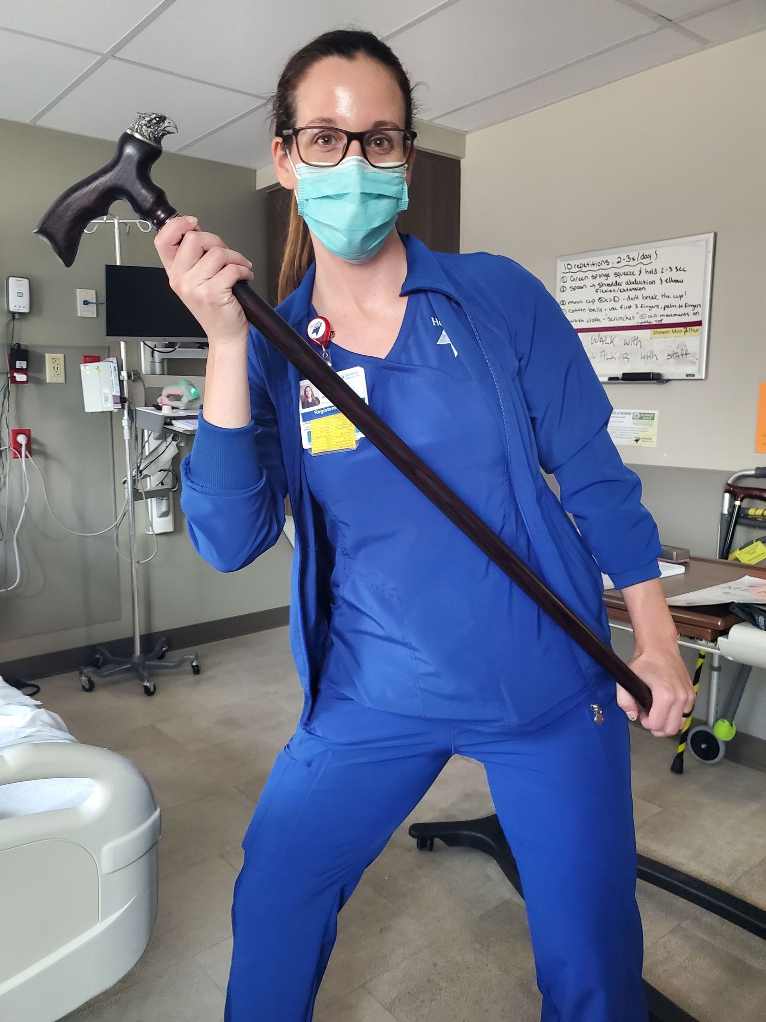 Healthcare worker in blue scrubs and surgical mask poses playfully with overhead examination light in medical room.