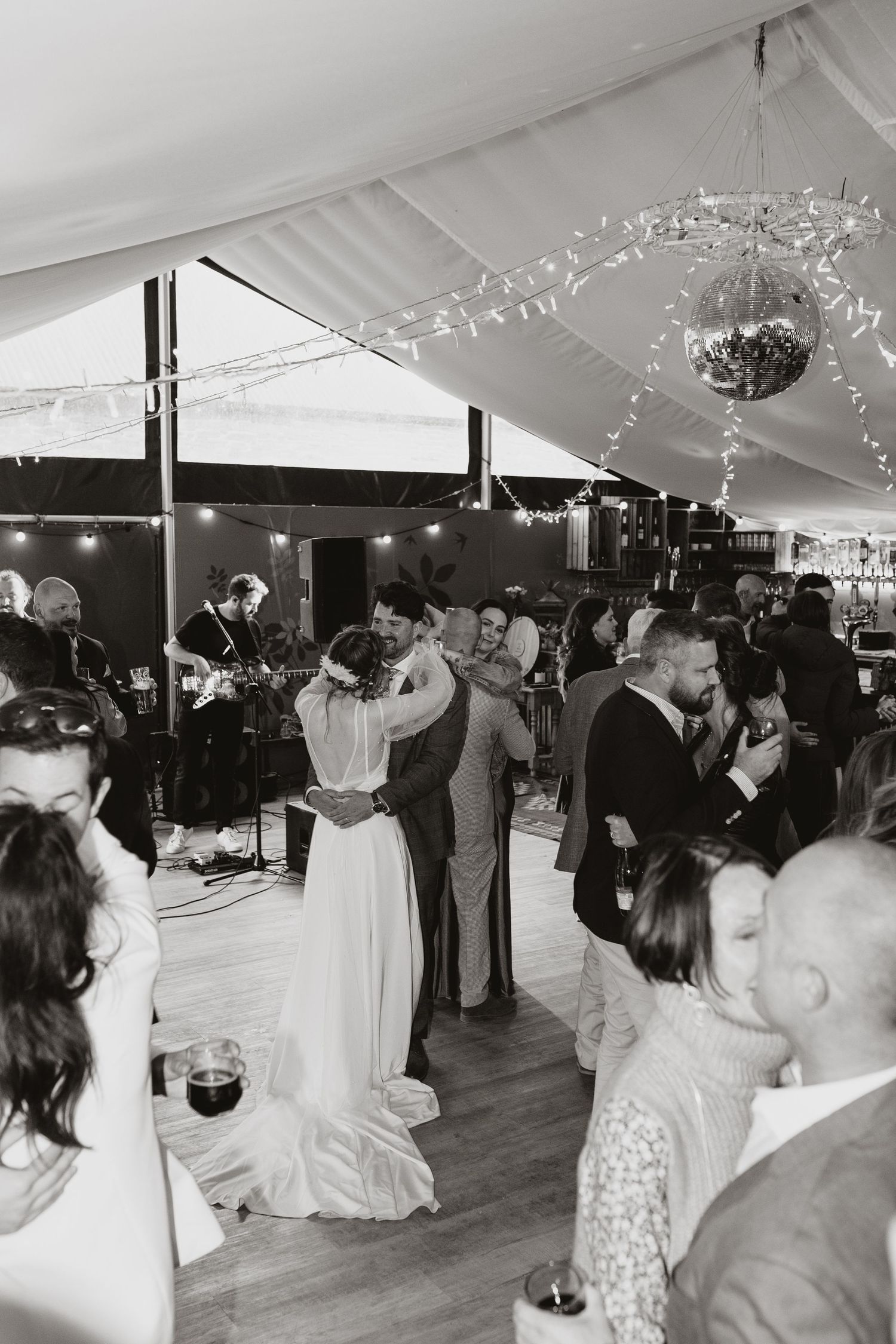 Black and white photo of couples dancing under a tent at an evening wedding reception with disco ball and string lights.