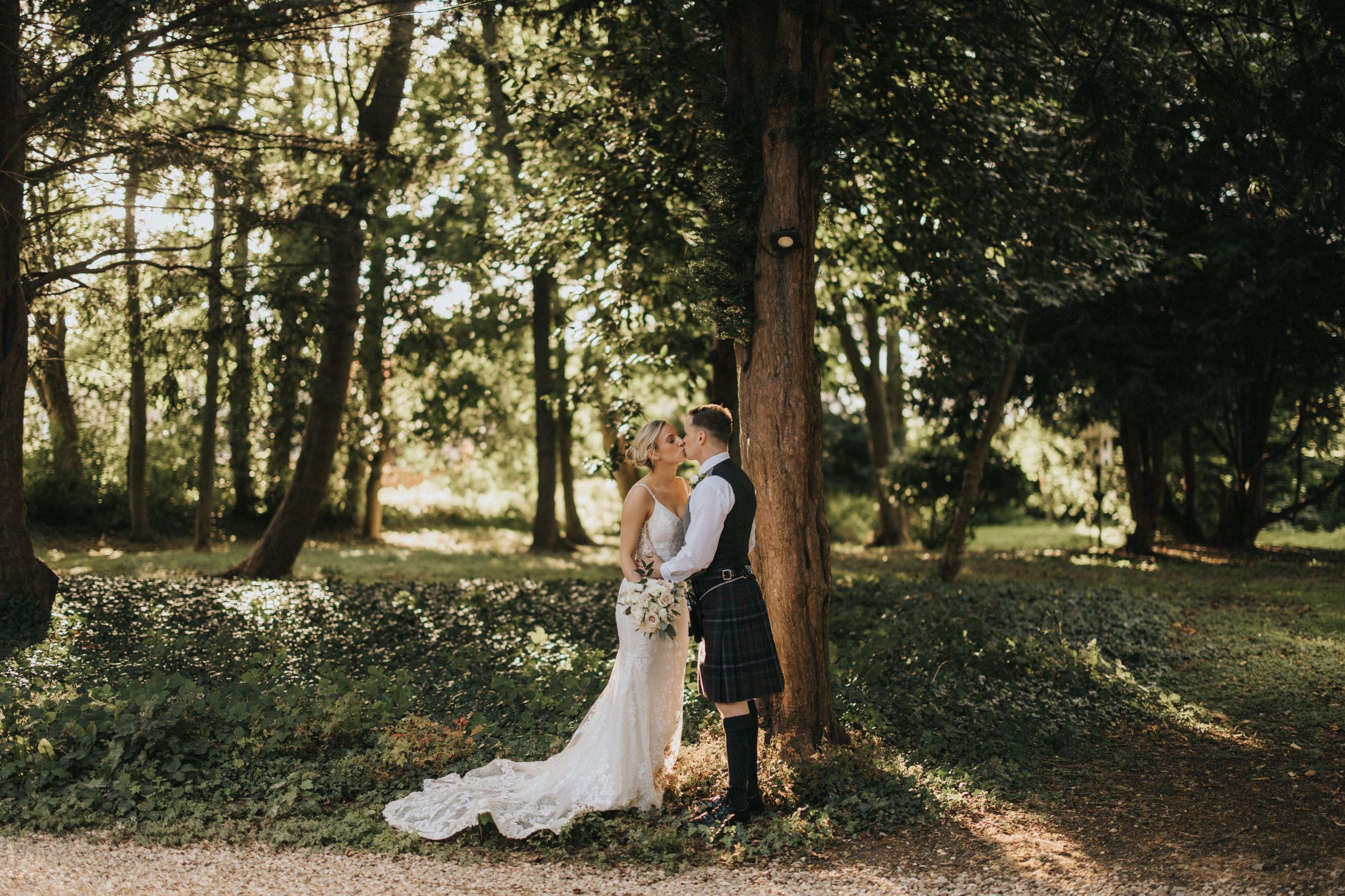 A couple in wedding attire embrace against a sunlit tree in a serene forest setting at Washingborough Hall - taken by Grimsby photographer.
